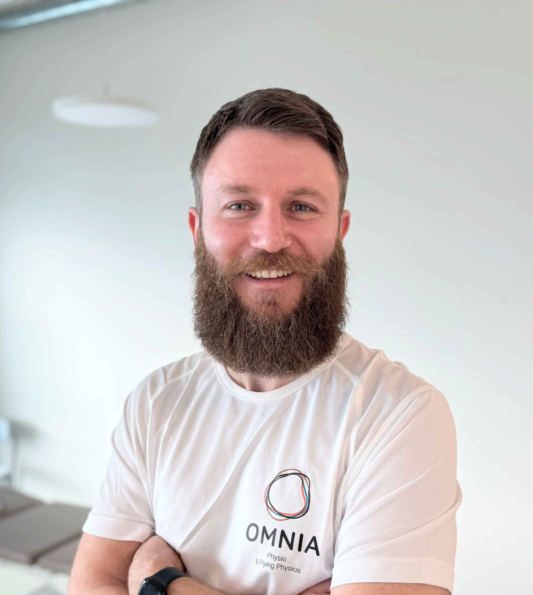 Mann mit blauen Augen und Bart trägt ein weißes T-Shirt mit dem OMNIA Logo und einem Geek-Schaltkreis auf der Brust, weil er an Physiotherapie und fliegenden Physiotherapeuten arbeitet.