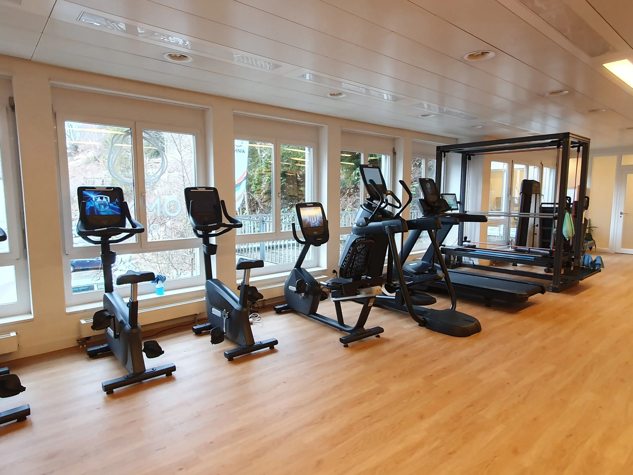 Innenaufnahme eines Fitnessstudios mit mehreren Cardio- und Kraftgeräten vor großen Fenstern.