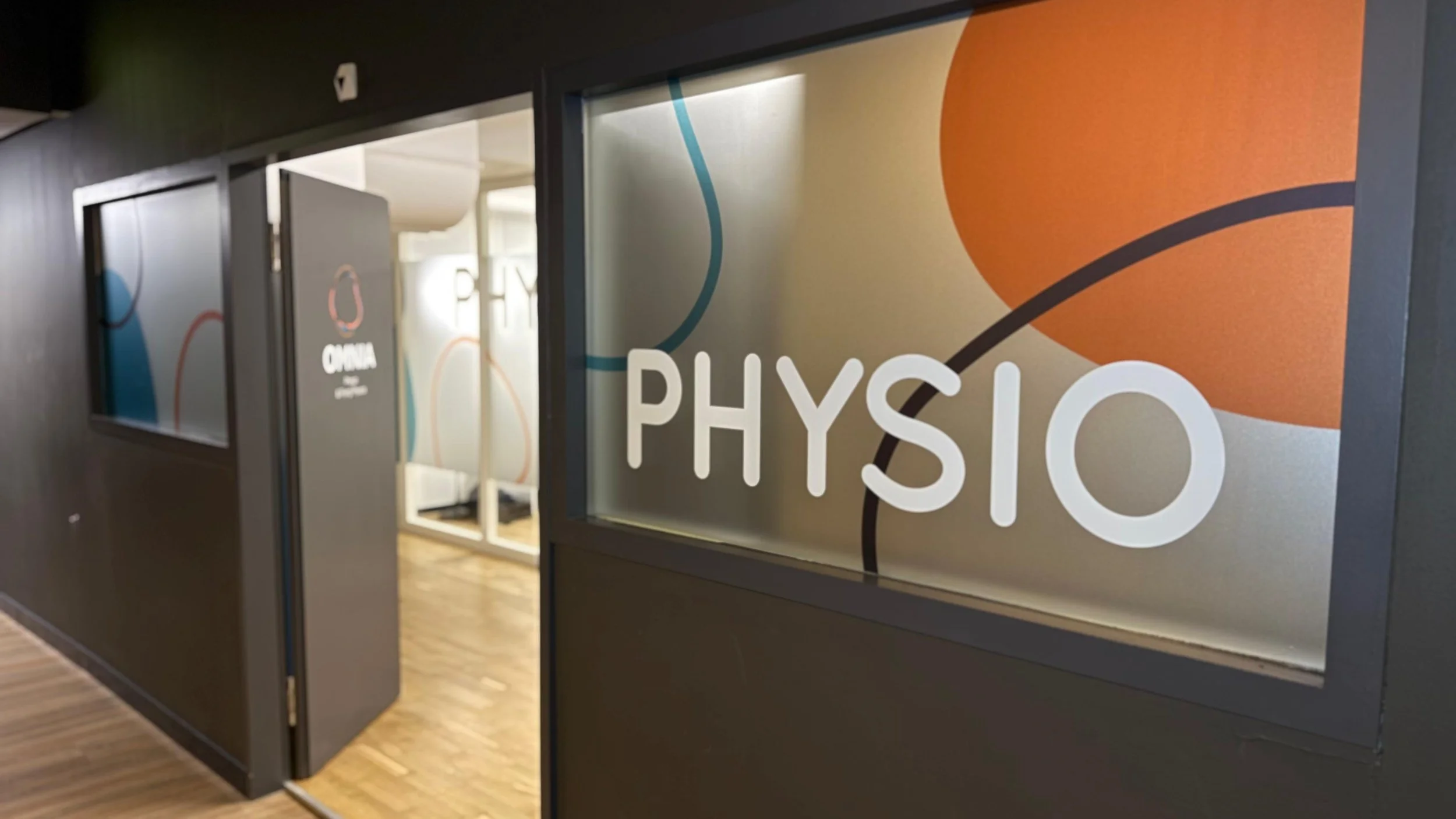 Innenansicht einer Physiotherapiepraxis mit einem Fenster, auf dem "PHYSIO" steht, und einem offenen Raum mit Therapieliegen.