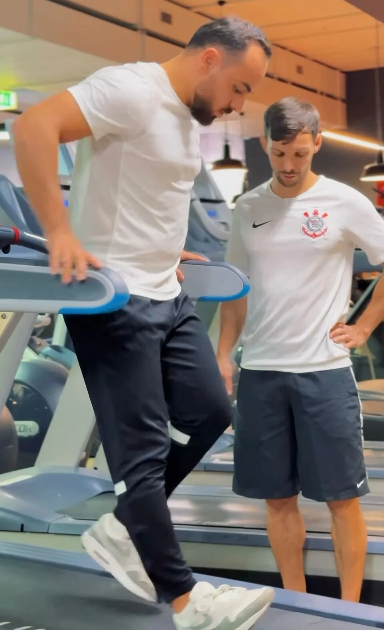 Zwei Männer im Fitnessstudio, einer benutzt das Laufband, der andere beobachtet ihn und trägt ein weißes T-Shirt mit einem Fußballverein-Logo.