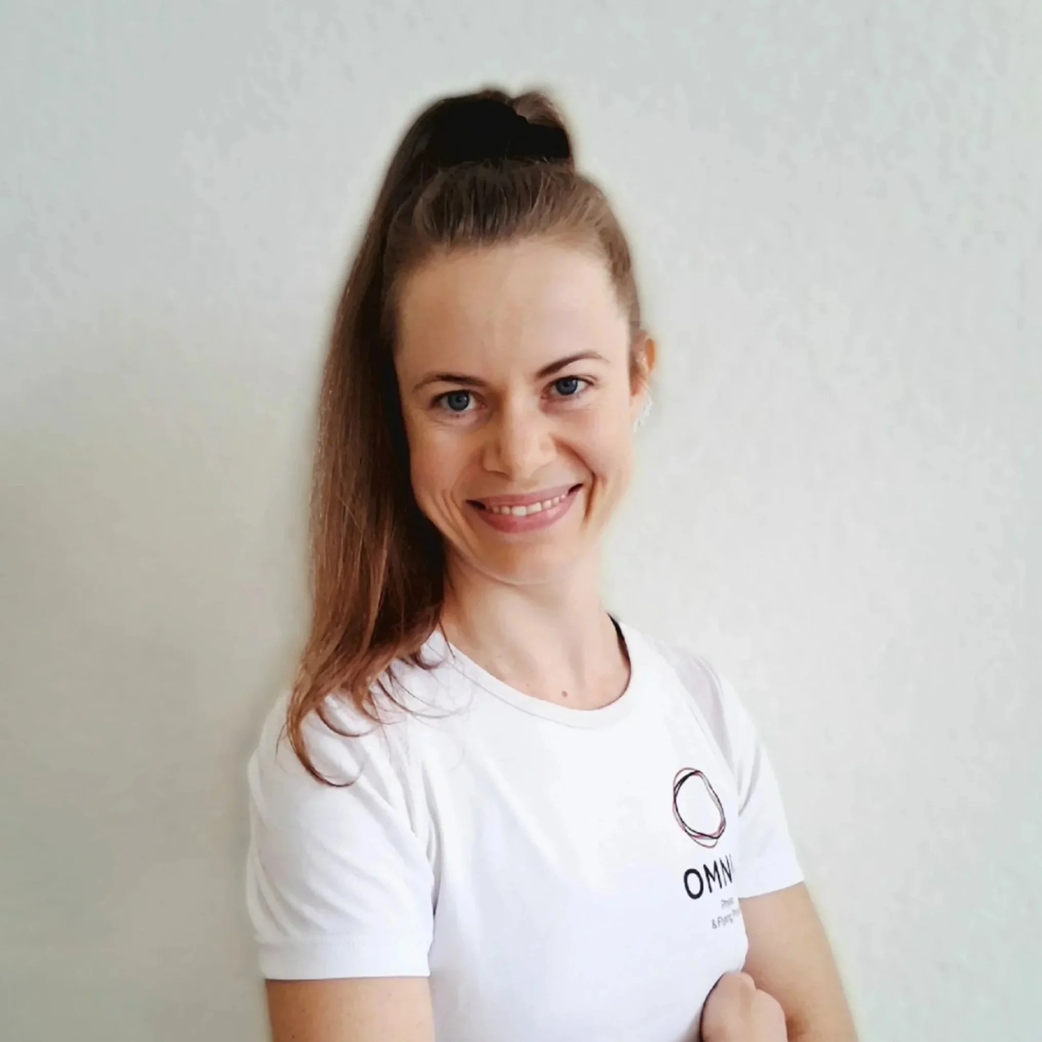 Junge Frau mit braunem Haar in einem hohen Pferdeschwanz, lächelnd vor weißer Wand, trägt weißes T-Shirt mit OMN Logo.