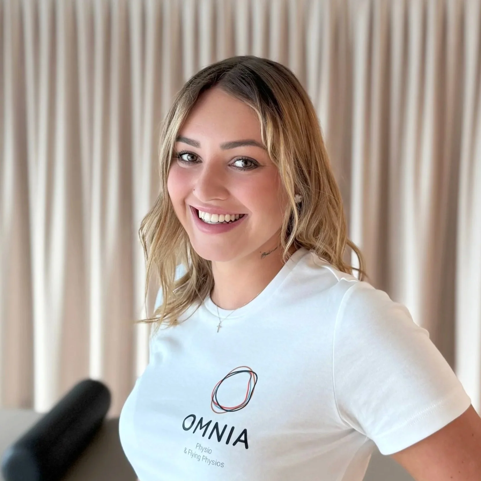 Lächelnde junge Frau mit braunen, gewellten Haaren, trägt ein weißes T-Shirt mit dem Logo 'OMNIA Physio & Flying Physios' vor einem beige-farbenen Vorhang.