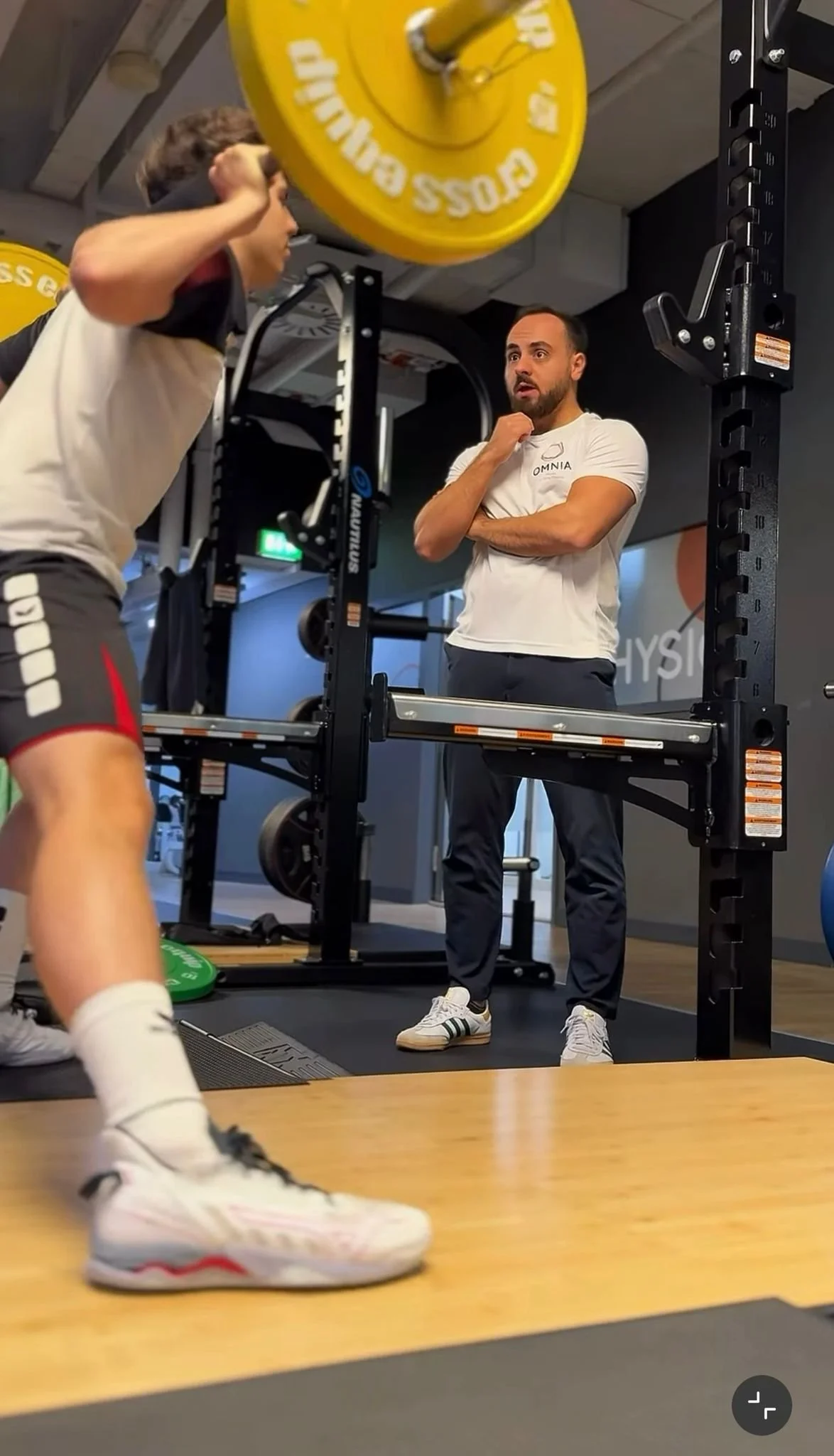 Ein Trainer beobachtet einen Jugendlichen beim Kniebeugen im Fitnessstudio, das Gewicht liegt auf den Schultern des Jugendlichen.