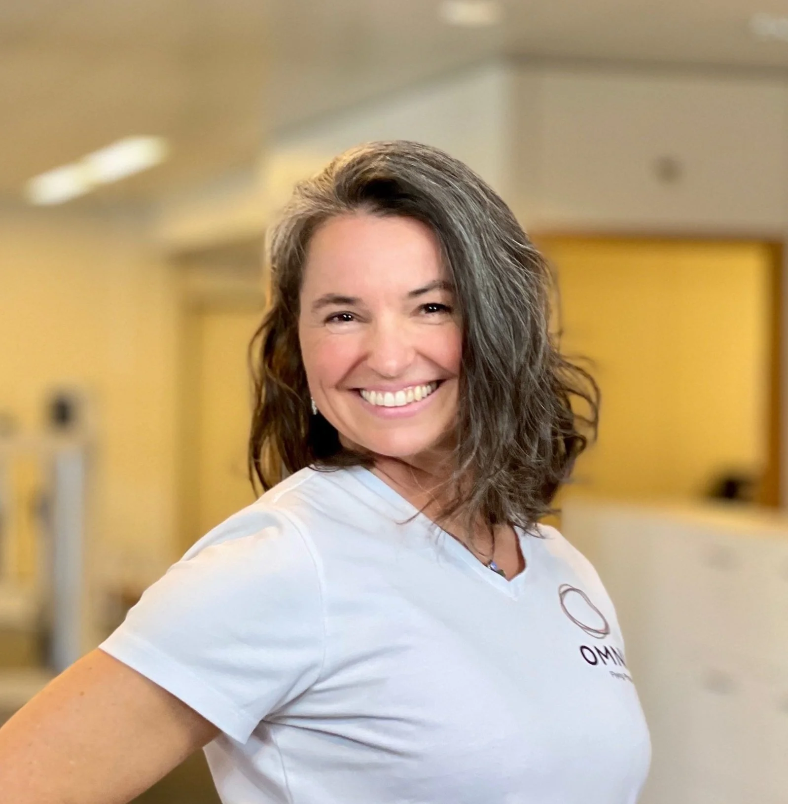 Lächelnde Frau mit braunen, gewellten Haaren trägt ein weißes T-Shirt mit OMNI-Logo im Hintergrund ist ein gelb gestrichener Raum.