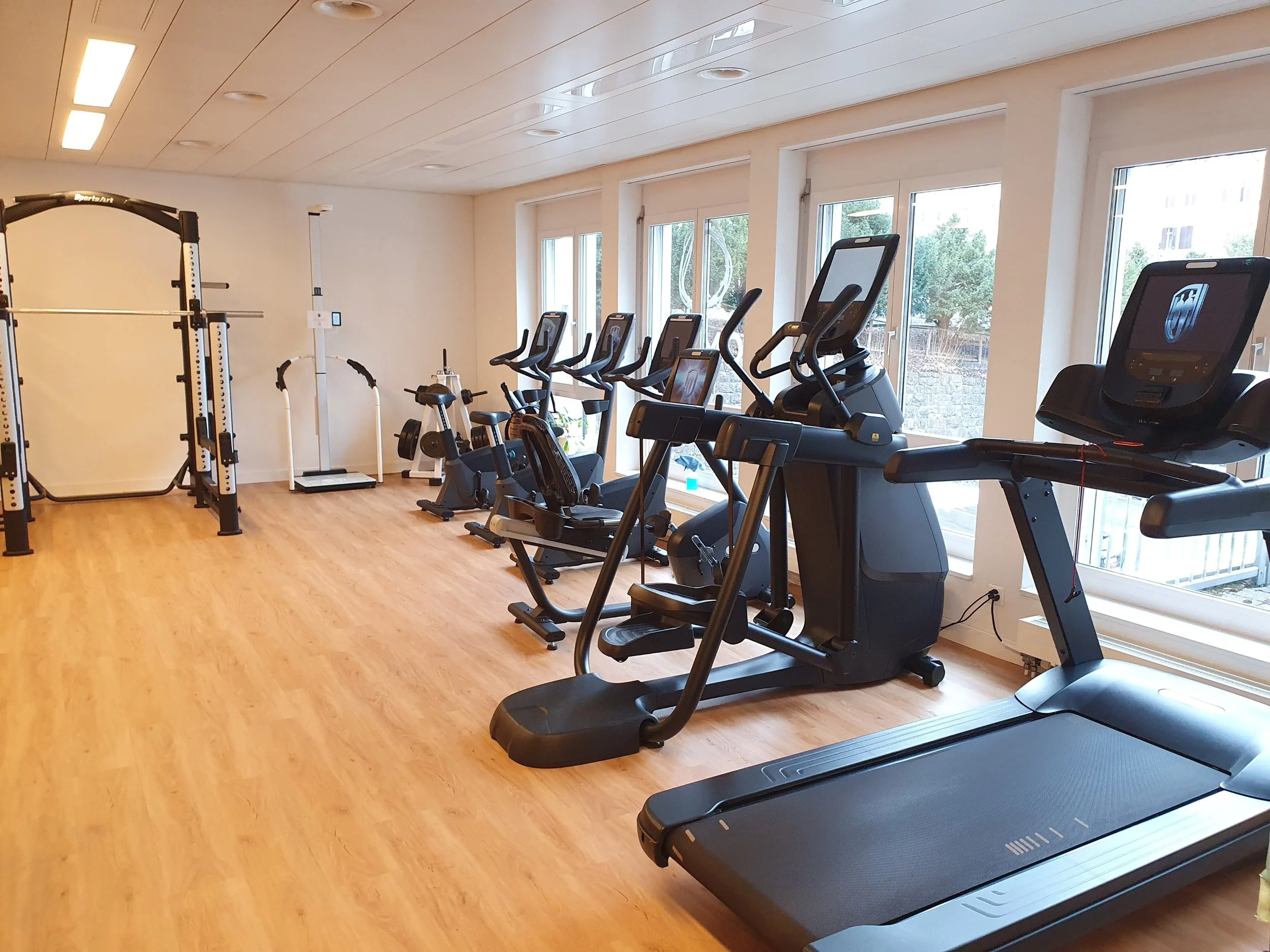 Fitnessstudio mit Laufband, Fahrradergometern, Hanteln und Kraftgeräten, großes Fenster an der Wandseite
