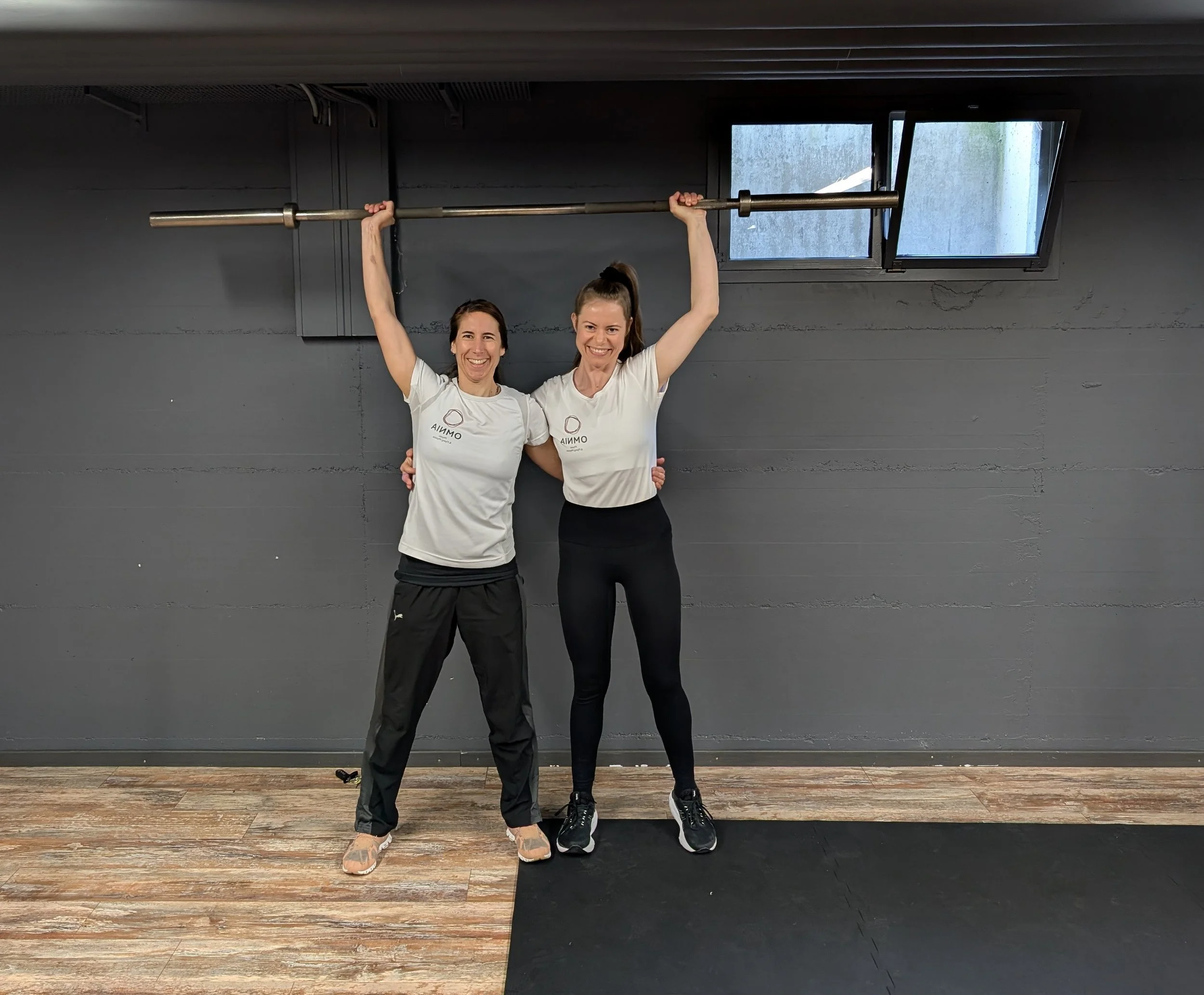 Zwei Frauen im Fitnessstudio, die eine Hantel Hebung machen, stehen nebeneinander, lächeln und tragen weiße T-Shirts.