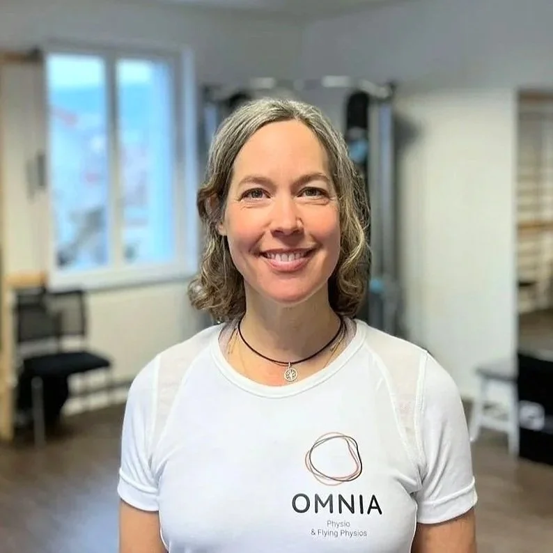 Lächelnde Frau mit grauen Haaren in einem Trainingsraum, trägt ein weißes T-Shirt mit dem Logo "OMNIA Physio & Flying Physios".