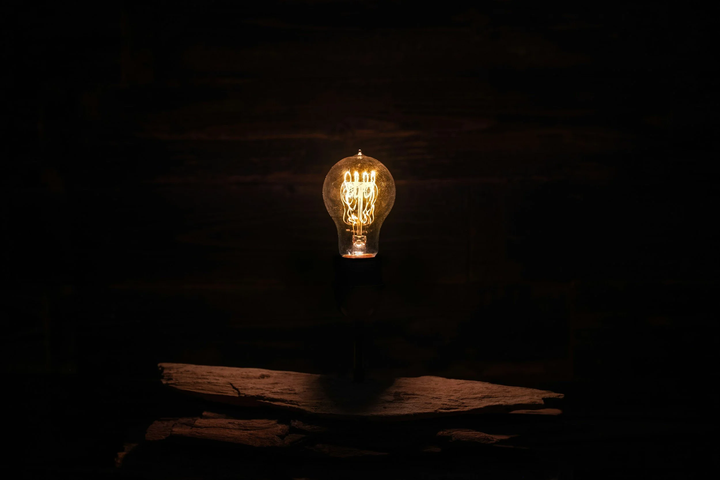 A glowing filament light bulb illuminated in a dark setting with a wooden surface beneath it.