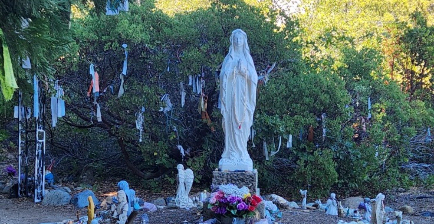 A religious shrine featuring a large white statue of the Virgin Mary surrounded by prayer ribbons, small statues, and flowers outdoors with greenery in the background.