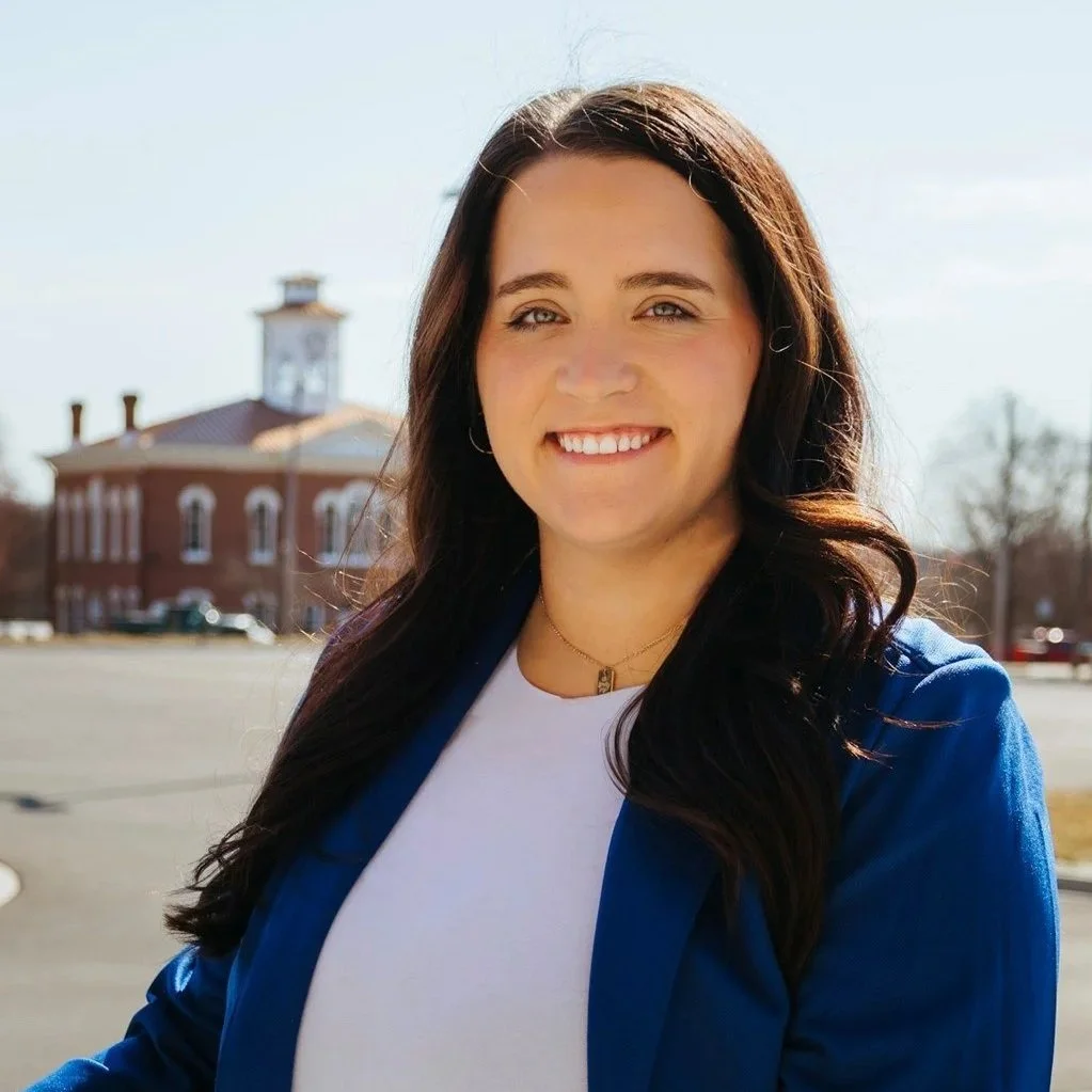 A headshot of Kassie Lex. Kassie has long dark hair and is smiling. She is wearing a white under a blue jacket.