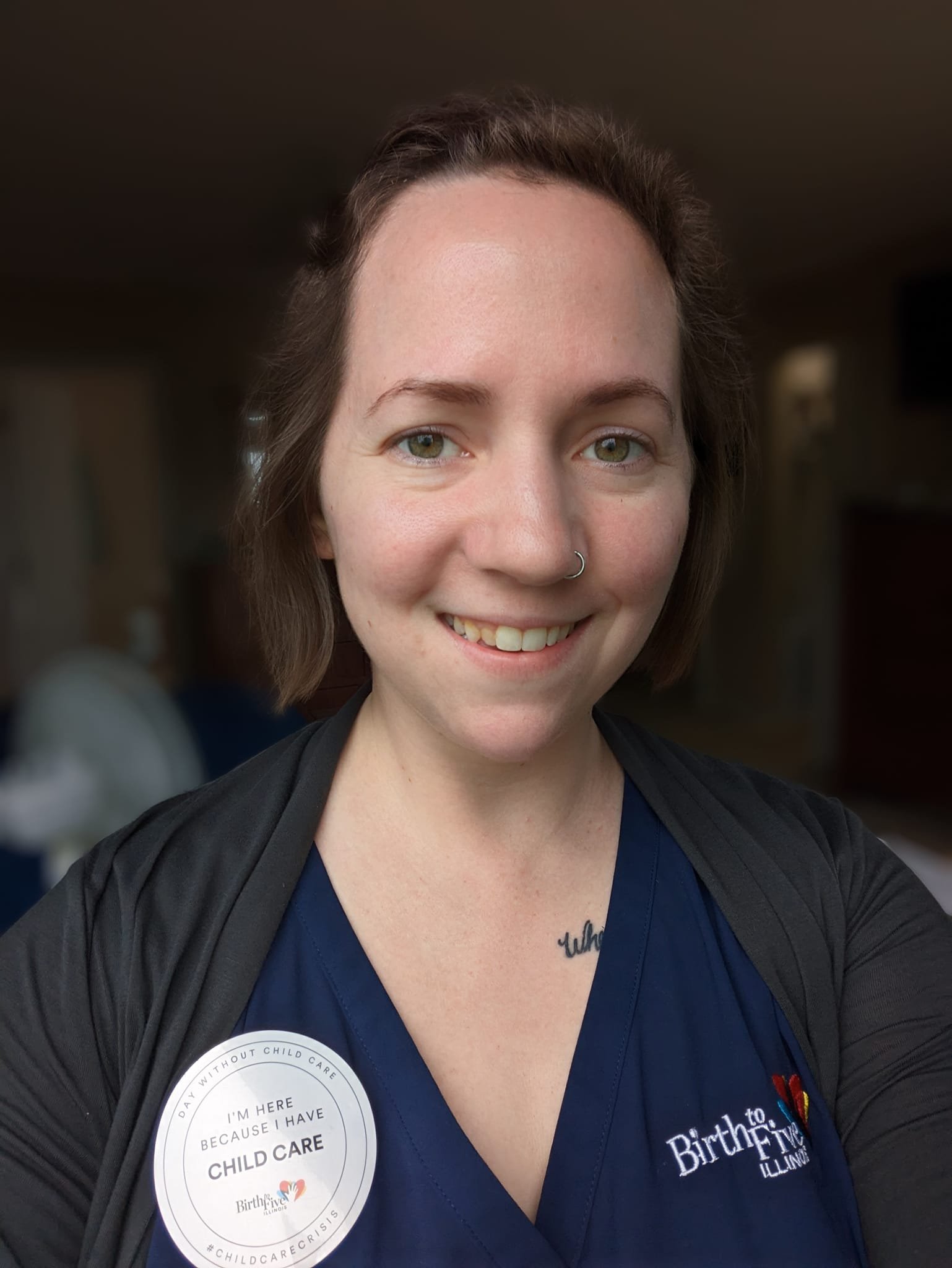 A picture of Gretchen Sprinkle smiling while wearing a button that says, “I’m here because I have child care.” The Birth to Five Illinois logo is underneath the sentence.