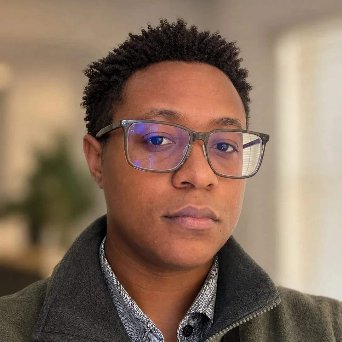 A headshot of Ty Patterson. They are wearing green glasses and a coat.