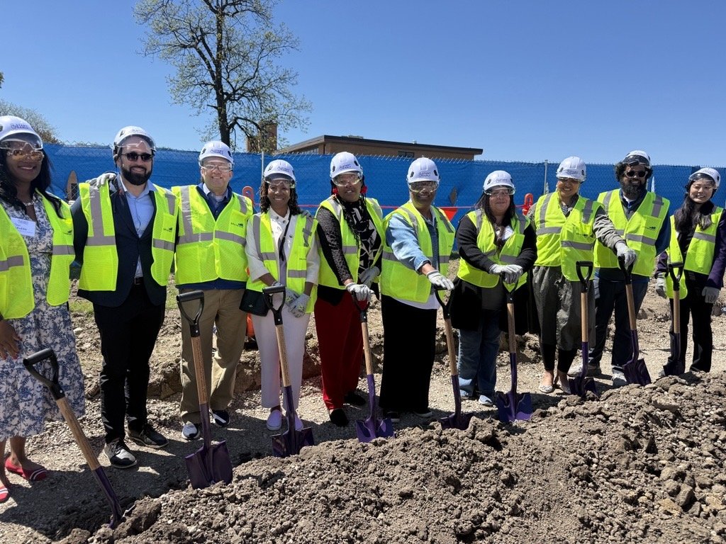 Picture of adults, all wearing hard hats, yellow vets, and holding shovels, at the ground breaking.