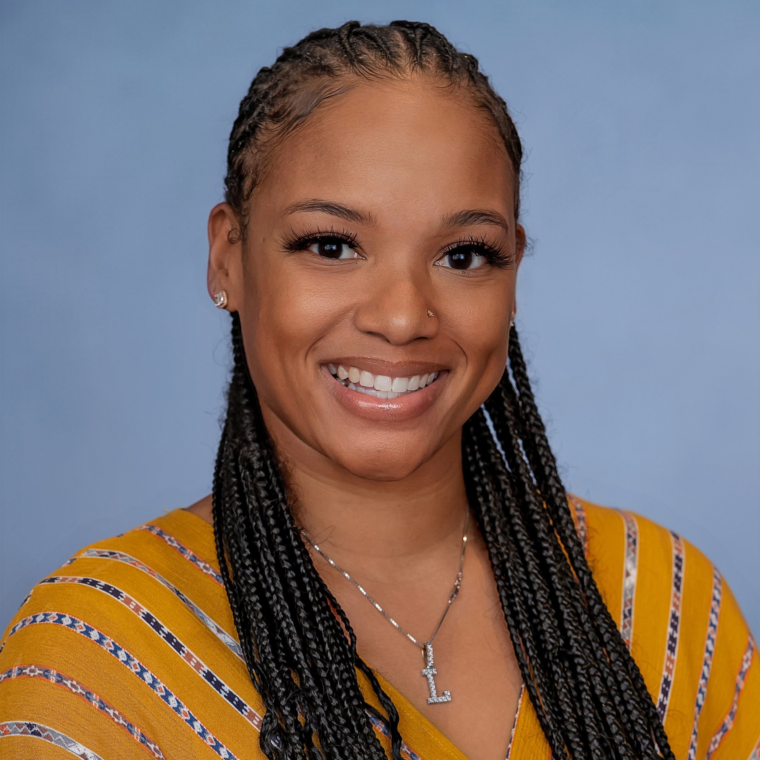 A headshot of Leah Armour. Leah has long dark braided hair and is smiling. Leah is wearing a orange patterned shirt with a necklace.