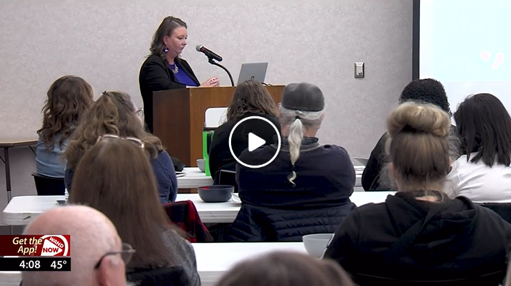 Screen grab of the TV  news segment. A person is standing at a podium in front of a room of people in chairs.