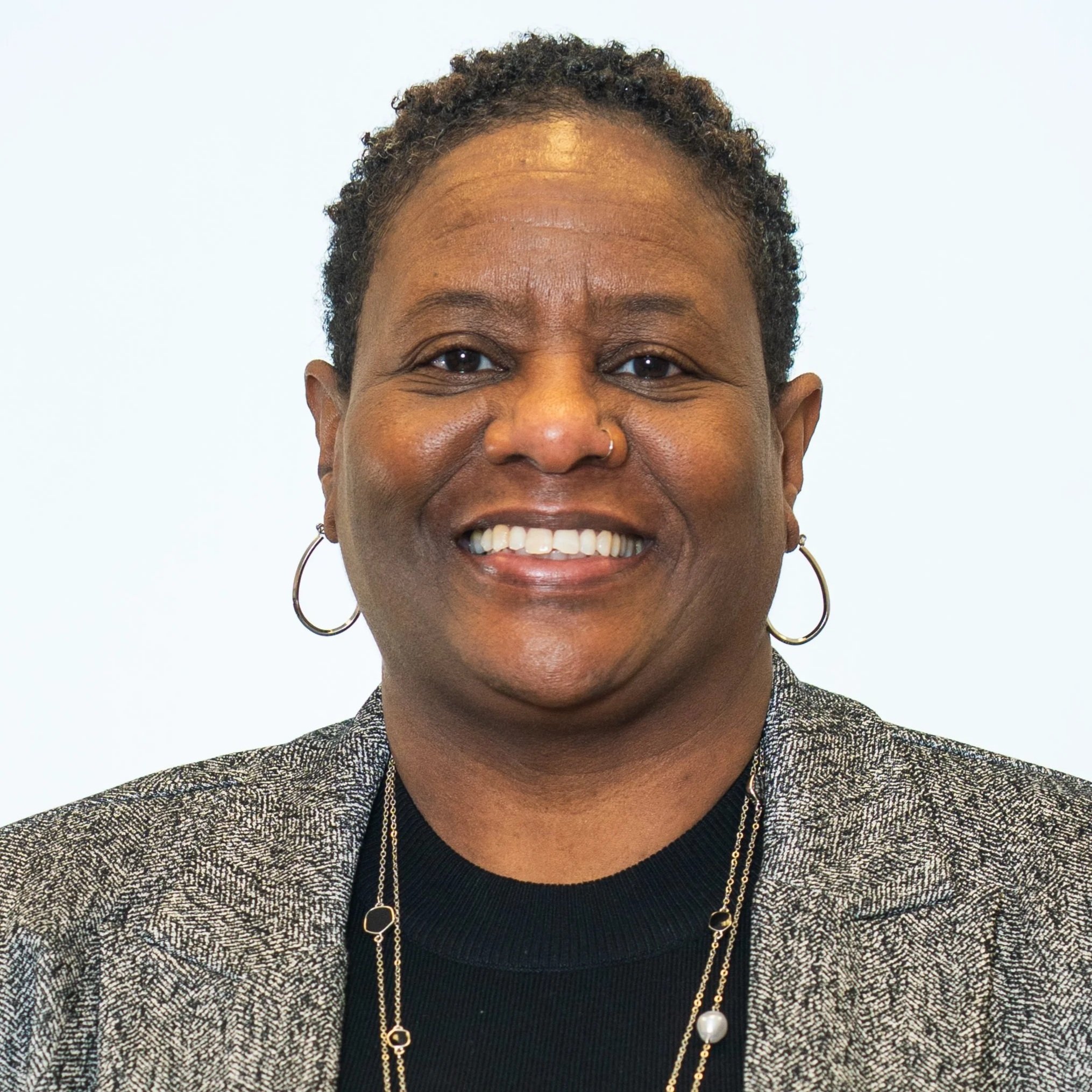 A headshot of Zena Madison. Zena is a dark-skinned person with short black hair. Zena is wearing a grey jacket over a black shirt with a gold necklace.