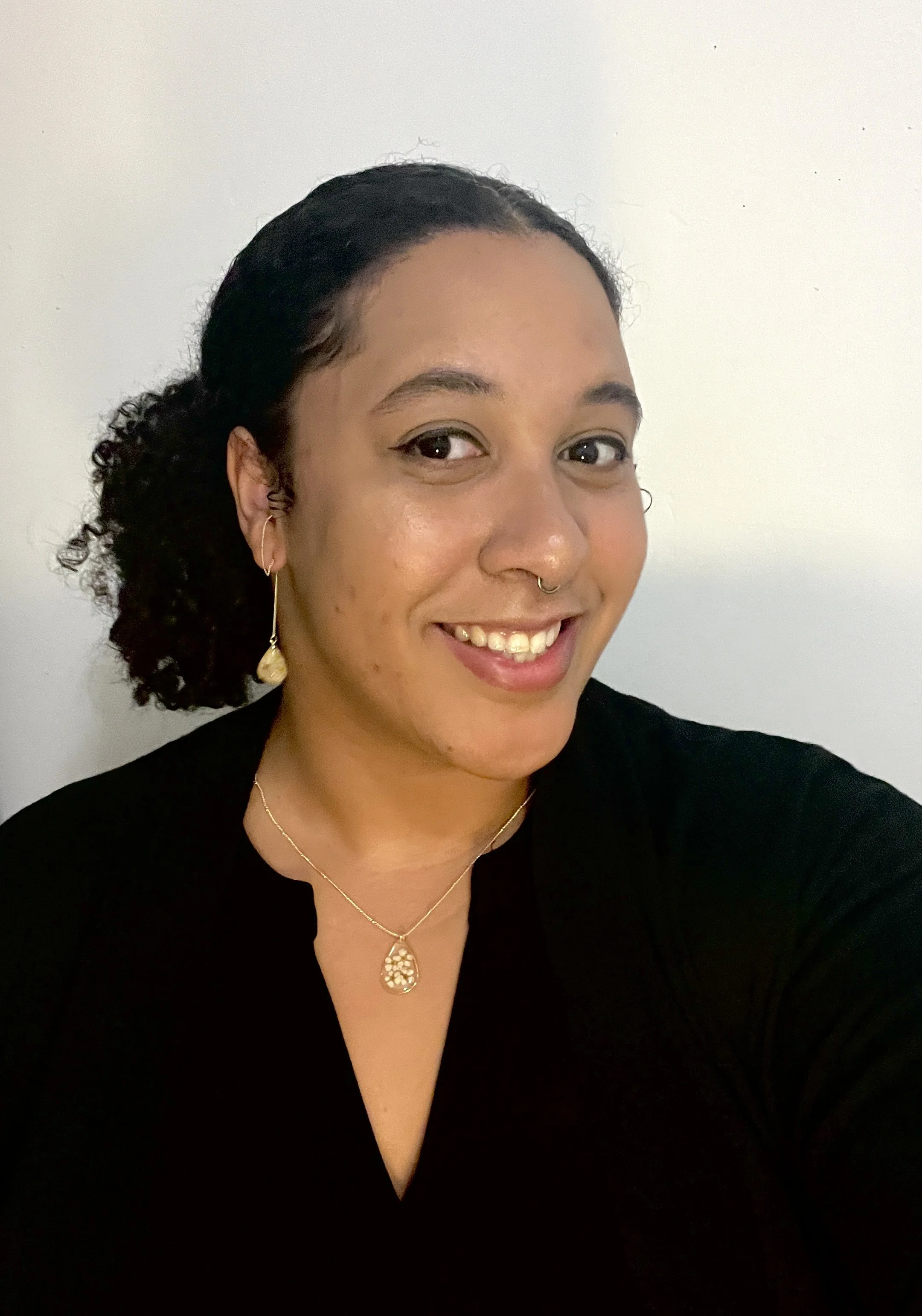 A woman with a medium-skin tone, and black curly hair in a ponytail. She has a small septum ring, long earrings, a black blouse, and a gold necklace.