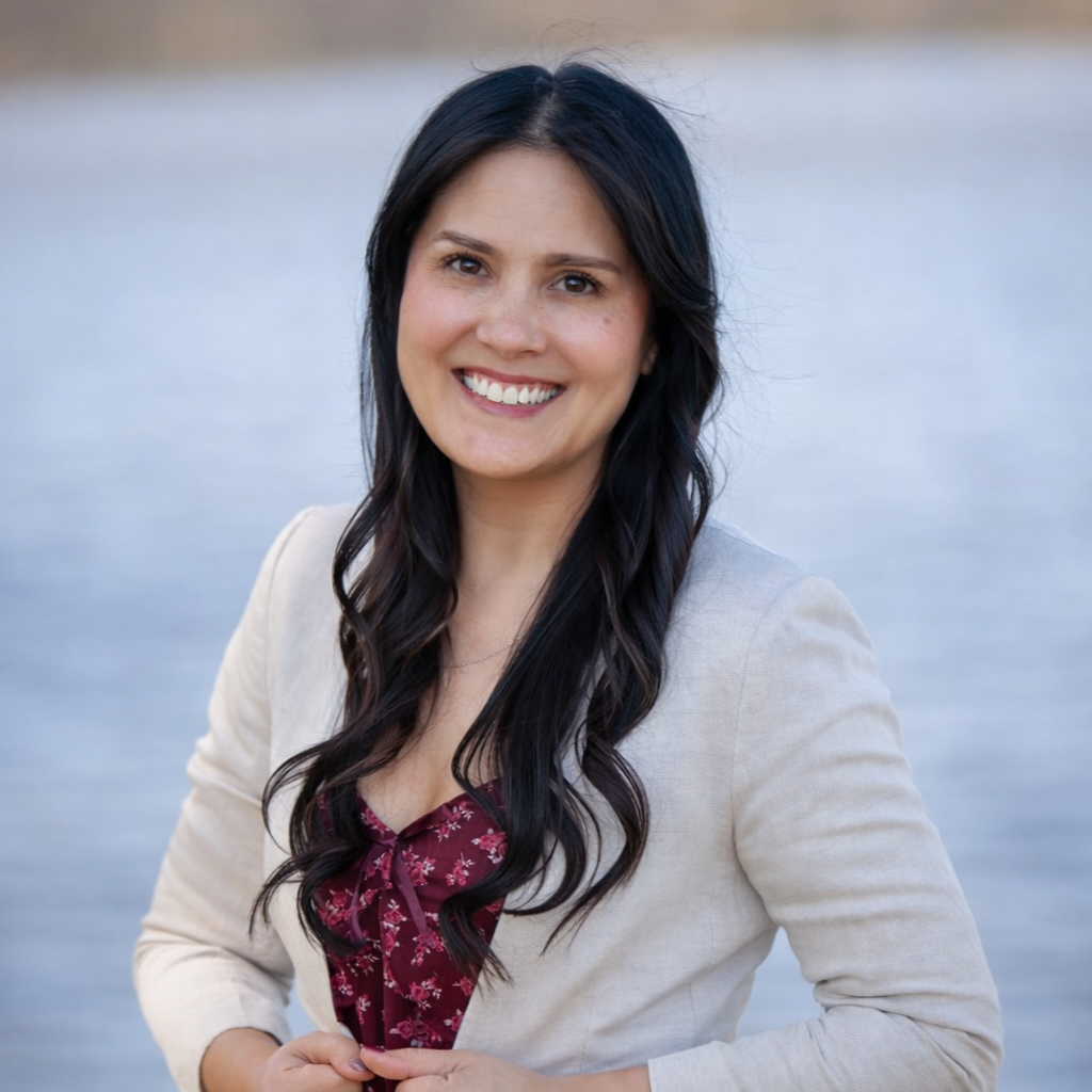 A headshot of Jessica Chin. Jessica has dark long hair and is smiling. Jessica is wearing a maroon floral top beneath a beige jacket.