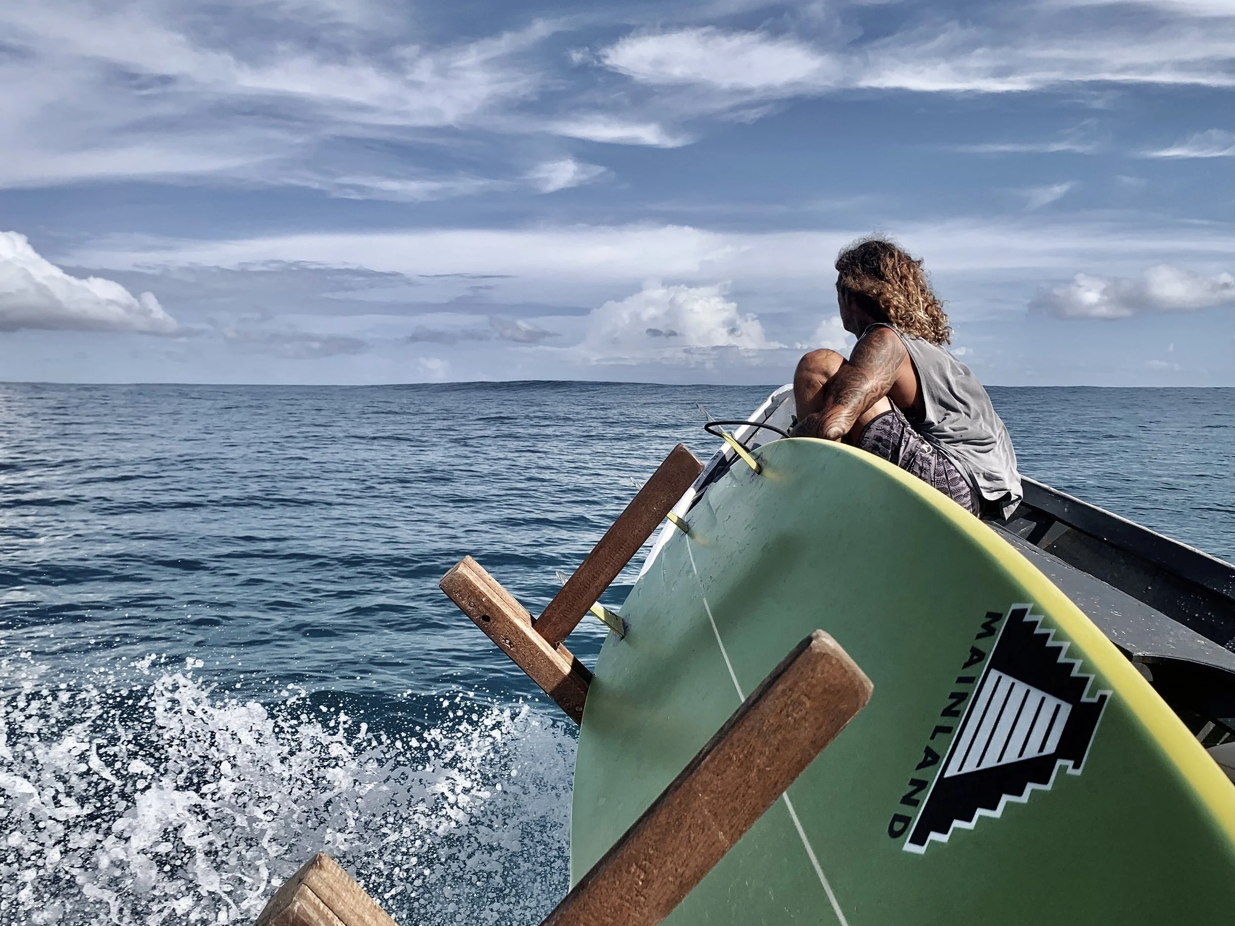Person with curly hair and tattoos on arm sitting on a boat with surfboard, looking at ocean and sky