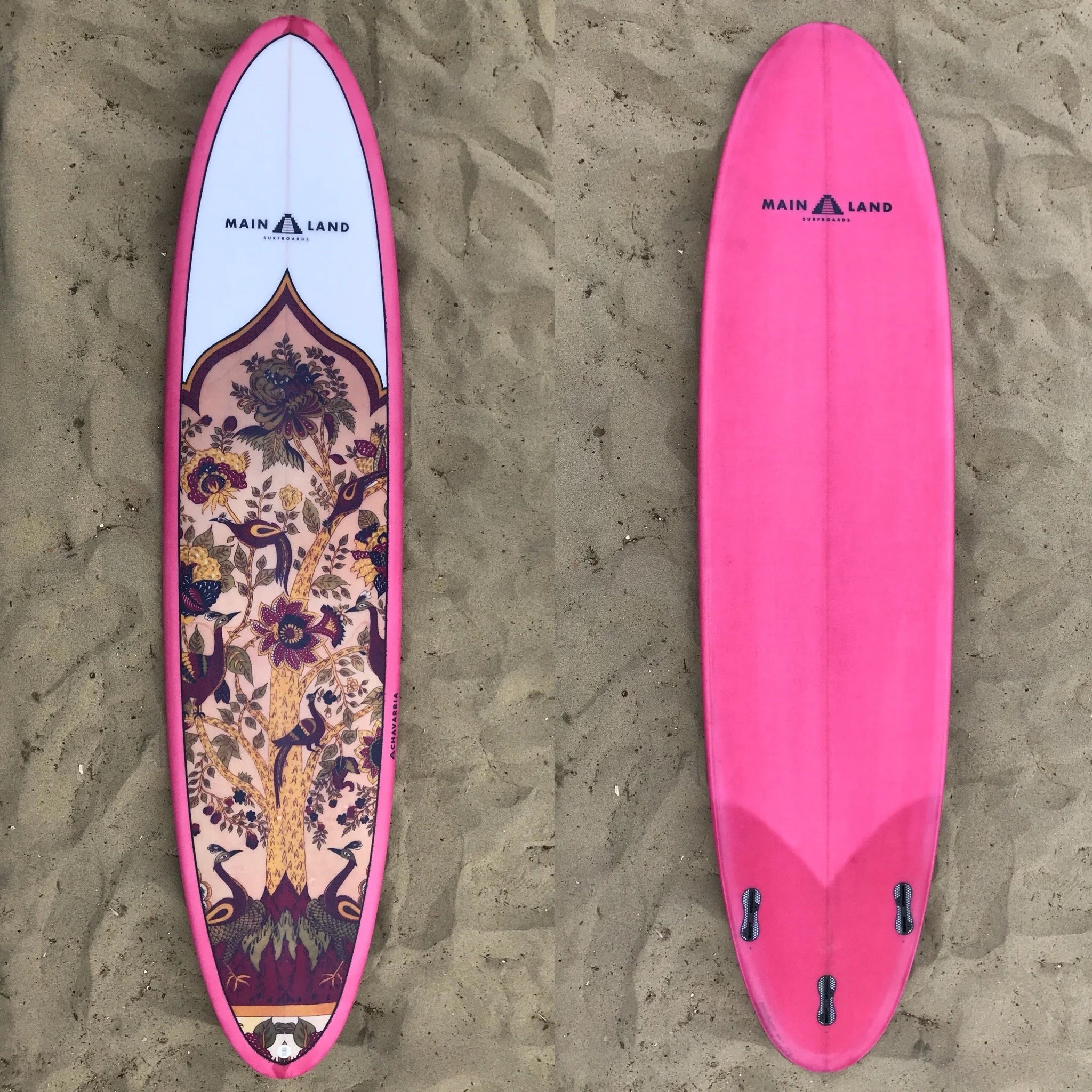 Two pink surfboards lying on the sand, one with a floral and bird design and the other plain pink.