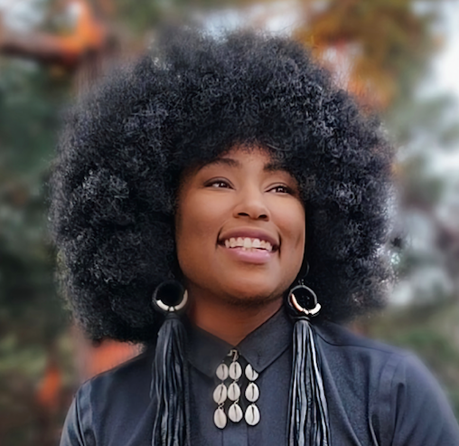 A smiling person with a large curly afro hairstyle wearing a navy jacket, shell earrings, and a shell necklace outdoors.