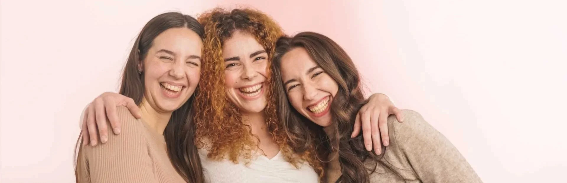 Three women with different hair colors and styles smiling and hugging each other against a light background.