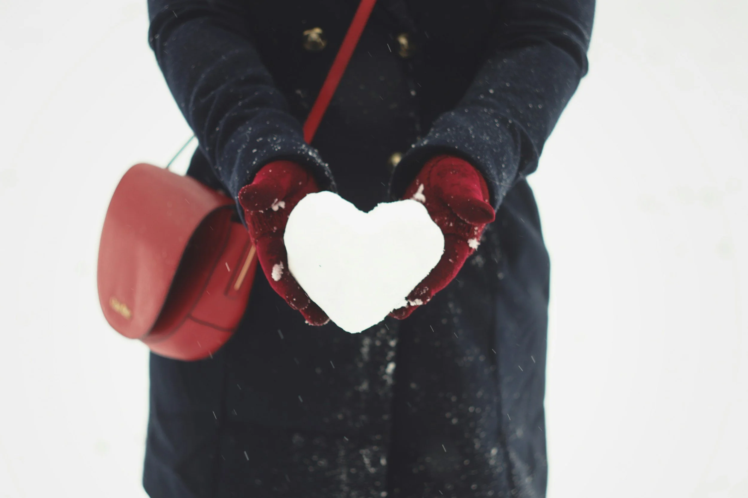 Person in winter clothing holding a snow heart, with a red bag hanging from their shoulder.