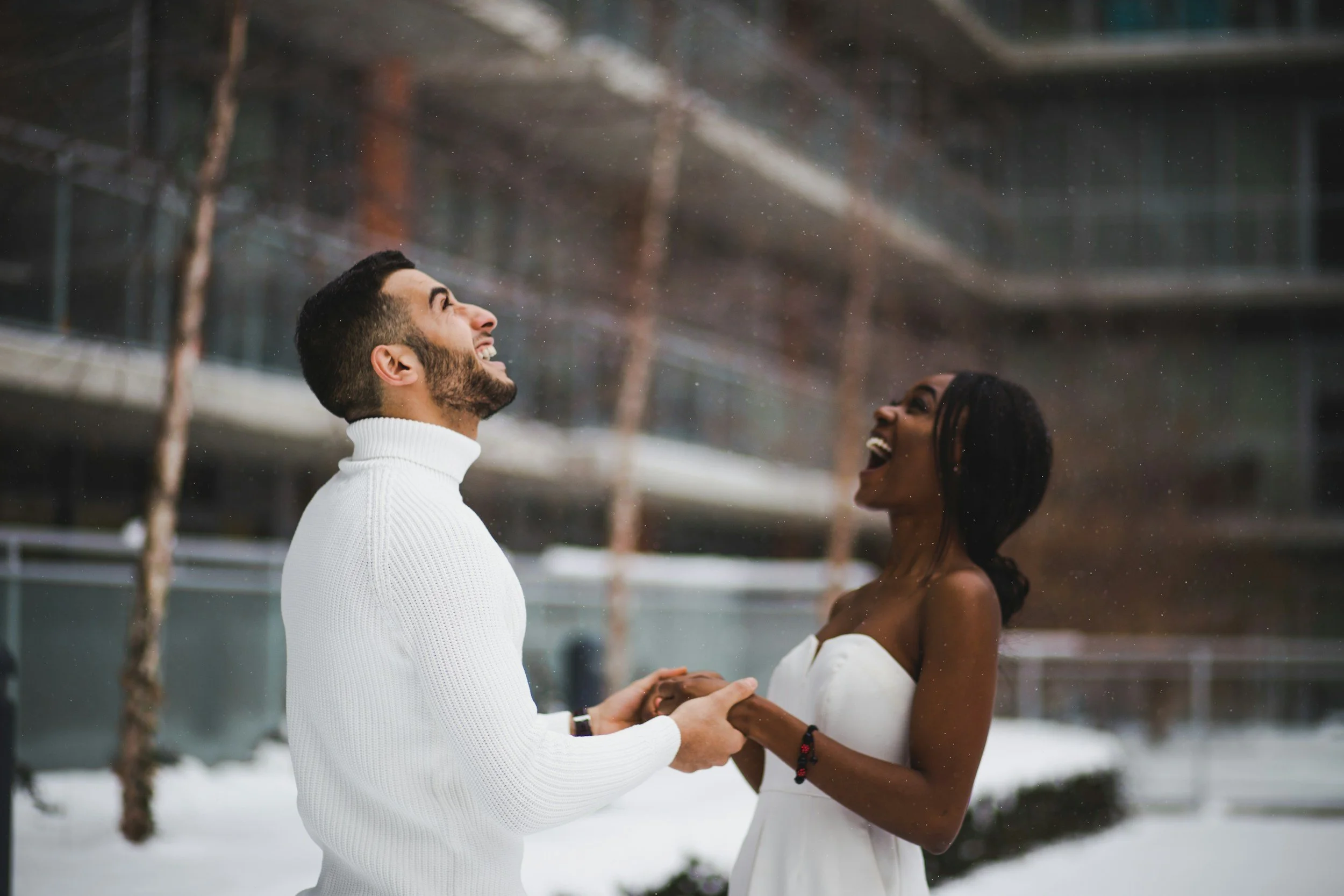 A joyful couple holding hands and laughing outdoors in a snowy setting, with a building and ropes in the background.