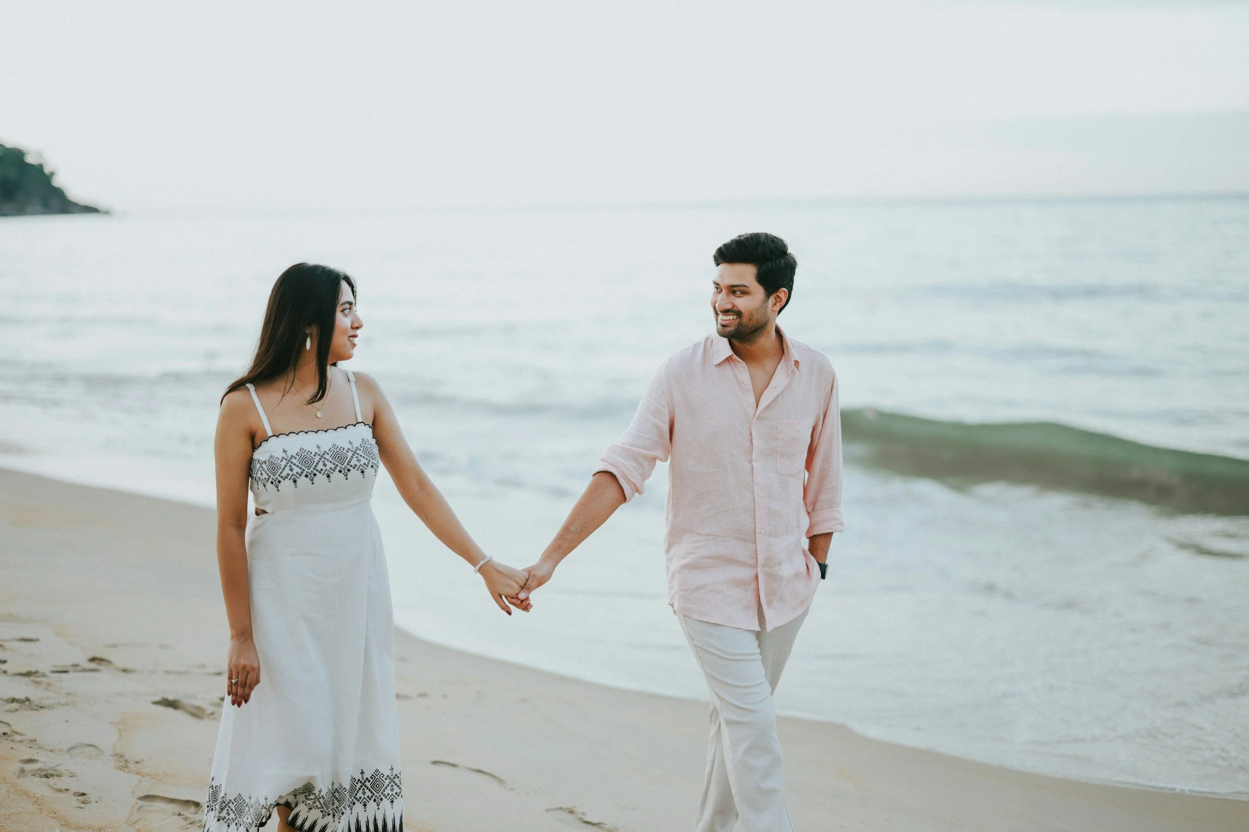 A couple holding hands and walking on a beach, facing each other and smiling, with the ocean in the background.