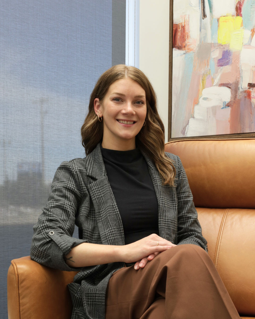 A woman with long wavy brown hair sitting on a tan leather couch, smiling at the camera, with an abstract painting on the wall behind her.