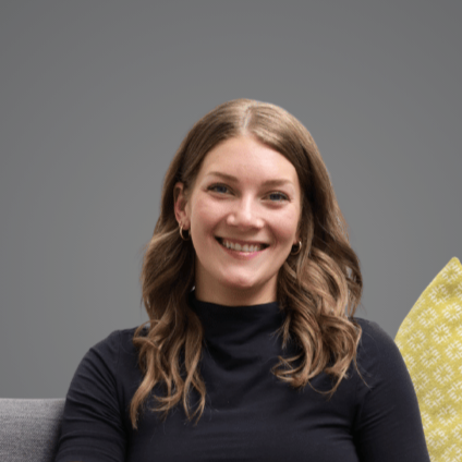 A woman with long, wavy brown hair smiling at the camera, wearing a black top, sitting on a gray chair against a plain gray background.