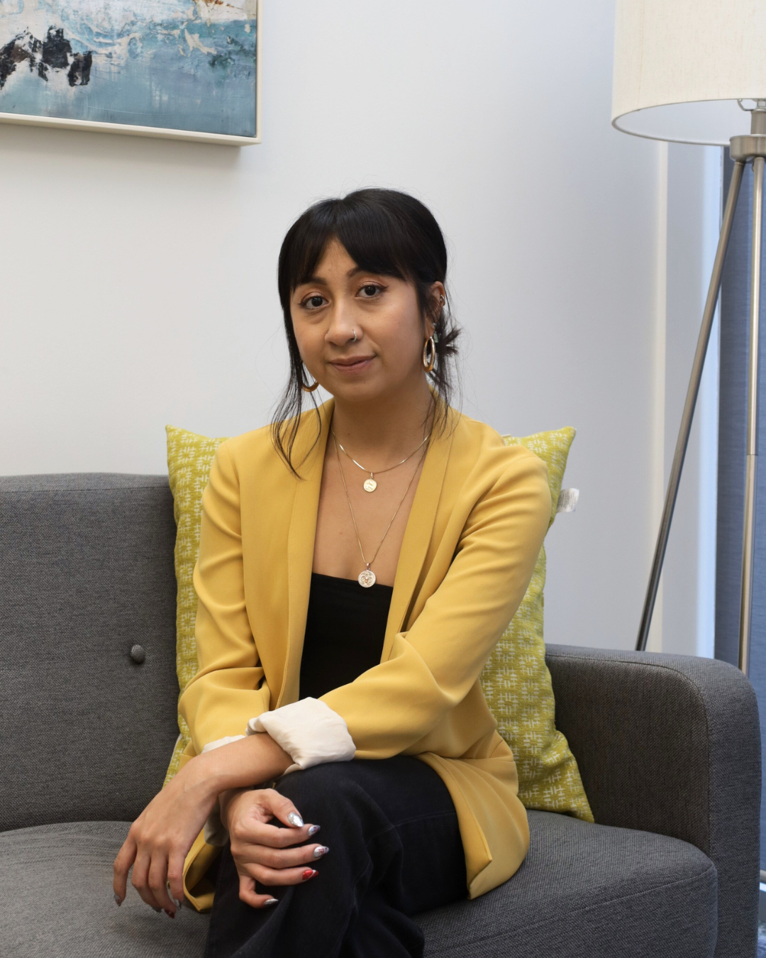A woman with short black hair, wearing a yellow blazer, black top, and jewelry, sitting on a gray sofa with a yellow cushion, in a room with a modern lamp and a painting on the wall.