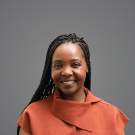 Portrait of a woman with braided hair wearing an orange top, smiling against a gray background.