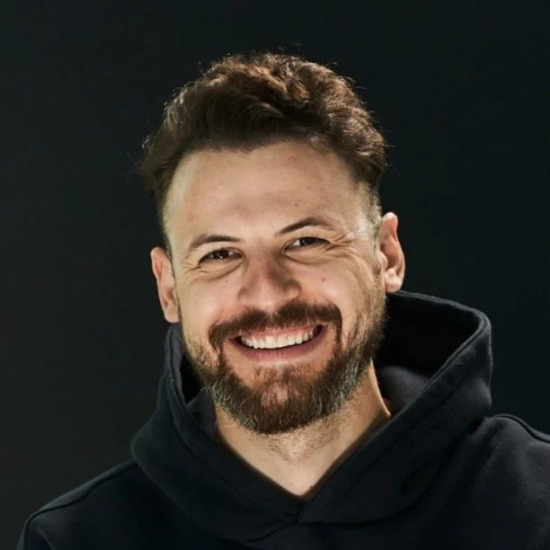 Photo of Michael Maximoff, a white man with brown curly hair and a brown beard smiling widely in front of a dark background.