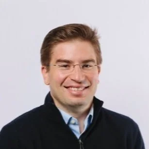 Photo of Chris (Christopher) Coad, a white man with brown hair wearing glasses with a thin transparent frame and smiling in front of a white background