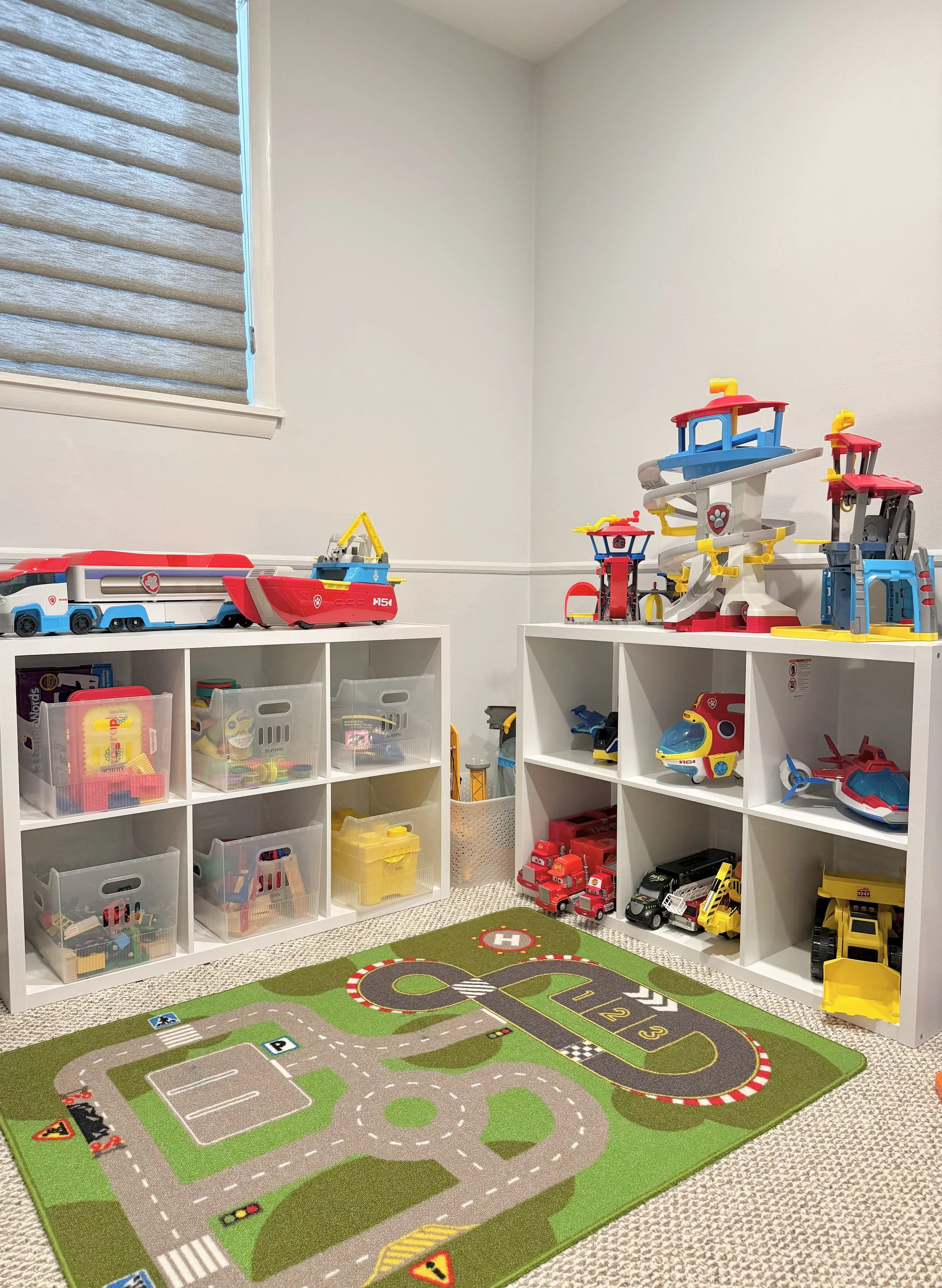 A playroom corner with white storage units holding toy vehicles and accessories, a colorful road-themed play mat on the floor, and a window with horizontal blinds on the left wall.