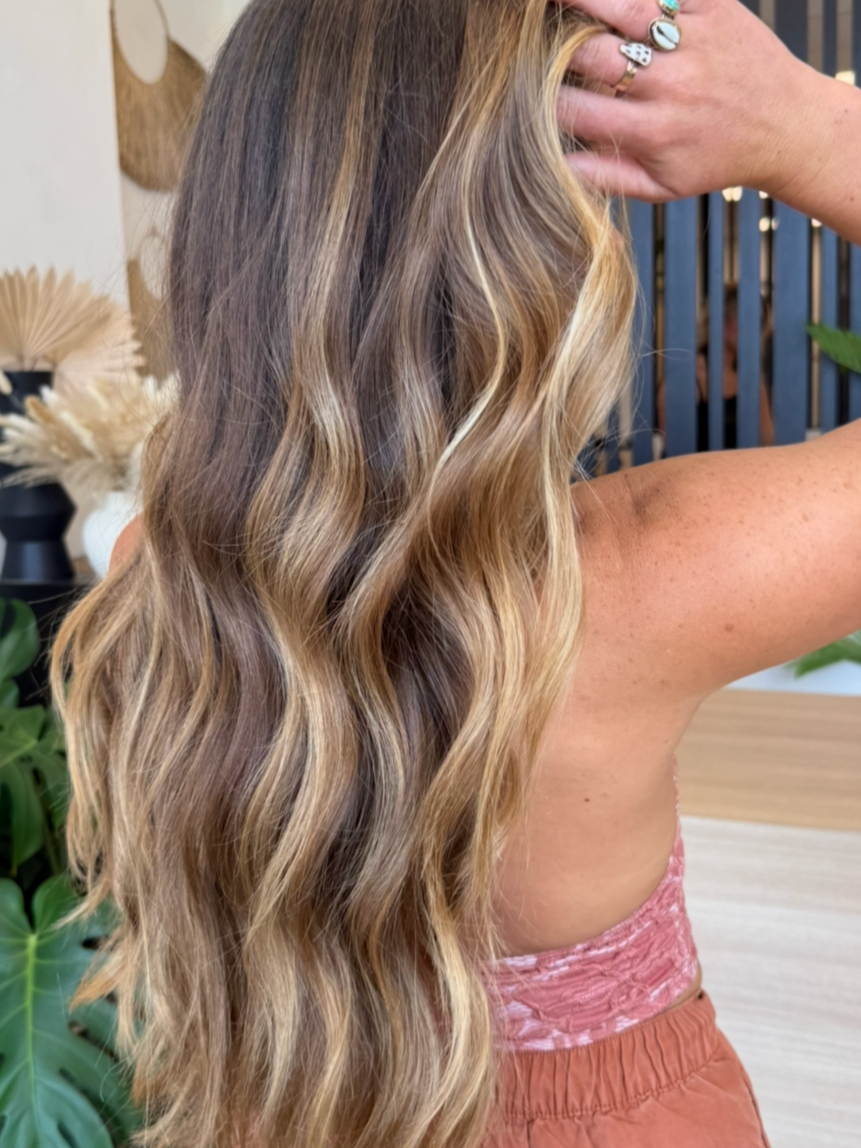 Close-up of a woman's long, wavy, blonde hair with highlights, held back by her hand, wearing rings, indoors with plants and decorative items in the background.