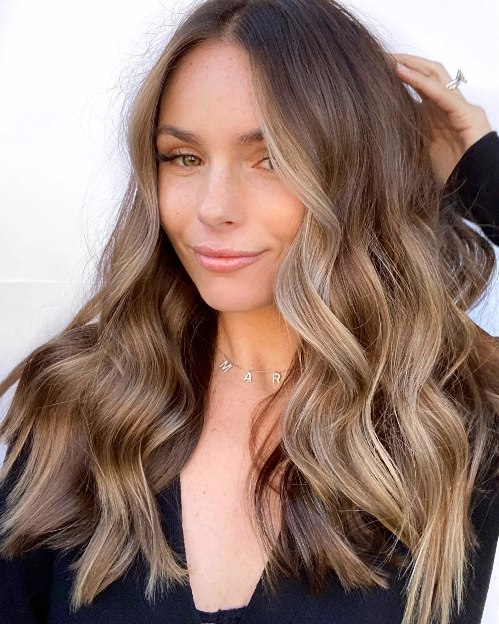Close-up of a woman with long, wavy, light brown hair with blonde highlights, smiling with one eye slightly closed, wearing a necklace and a black top.
