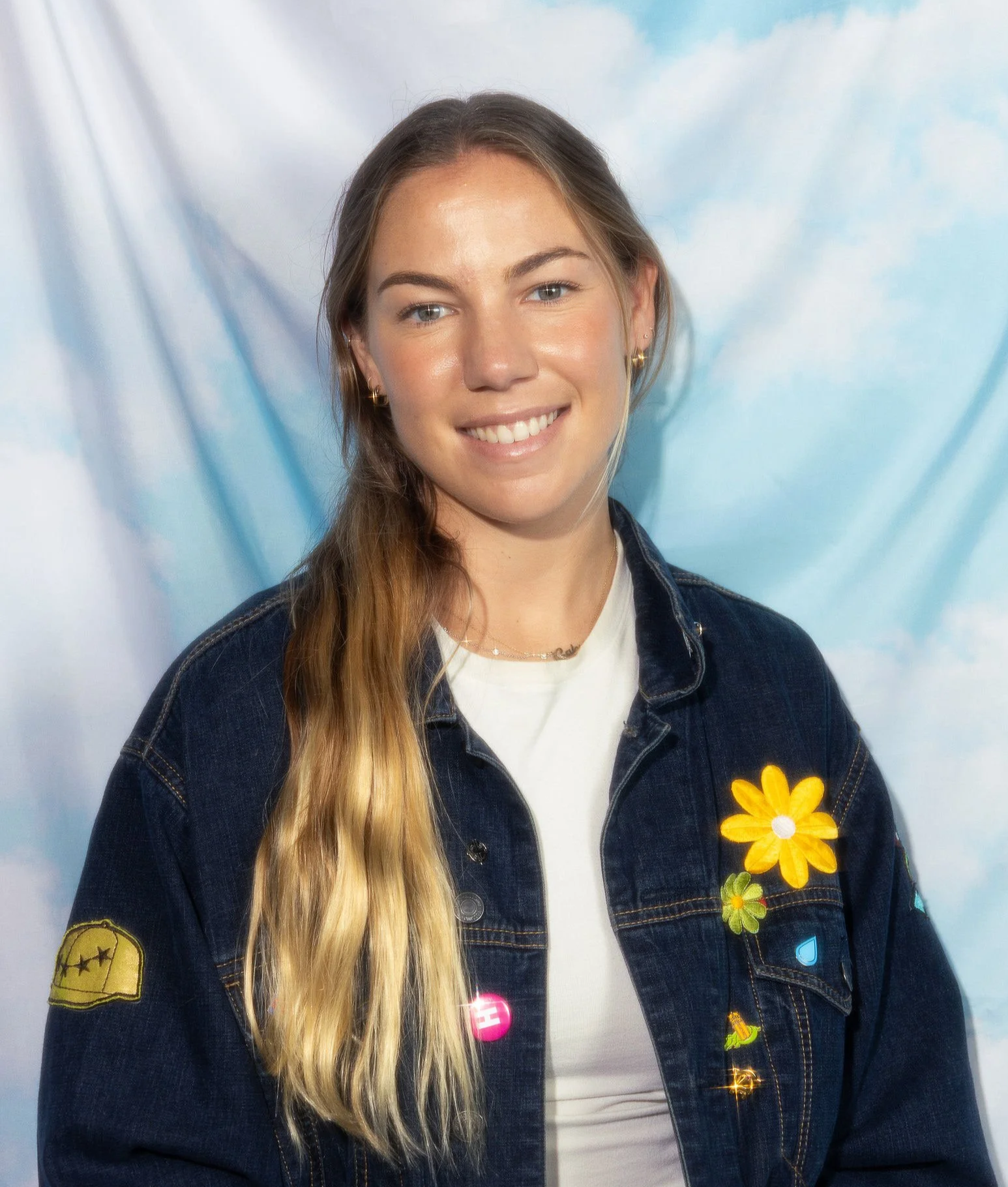 Young woman with long, wavy blonde hair, smiling, wearing a denim jacket with colorful patches and a yellow flower on the lapel, against a light blue and white cloudy background.