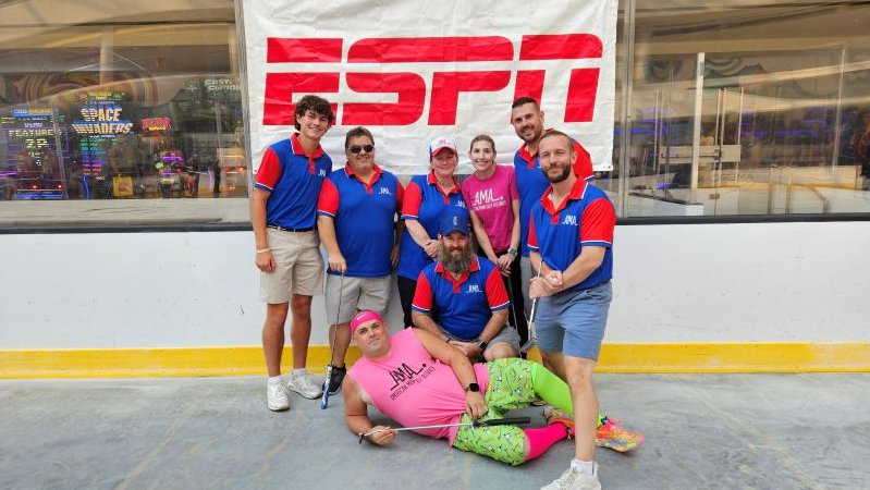 8 AMA minigolfers, 6 male and 2 female, posing in front of an ESPN sign in a de-iced hockey rink.  5 of the males and 1 female are wearing a blue and red AMA polo and  1 male and 1 female are wearing pink AMA shirts.