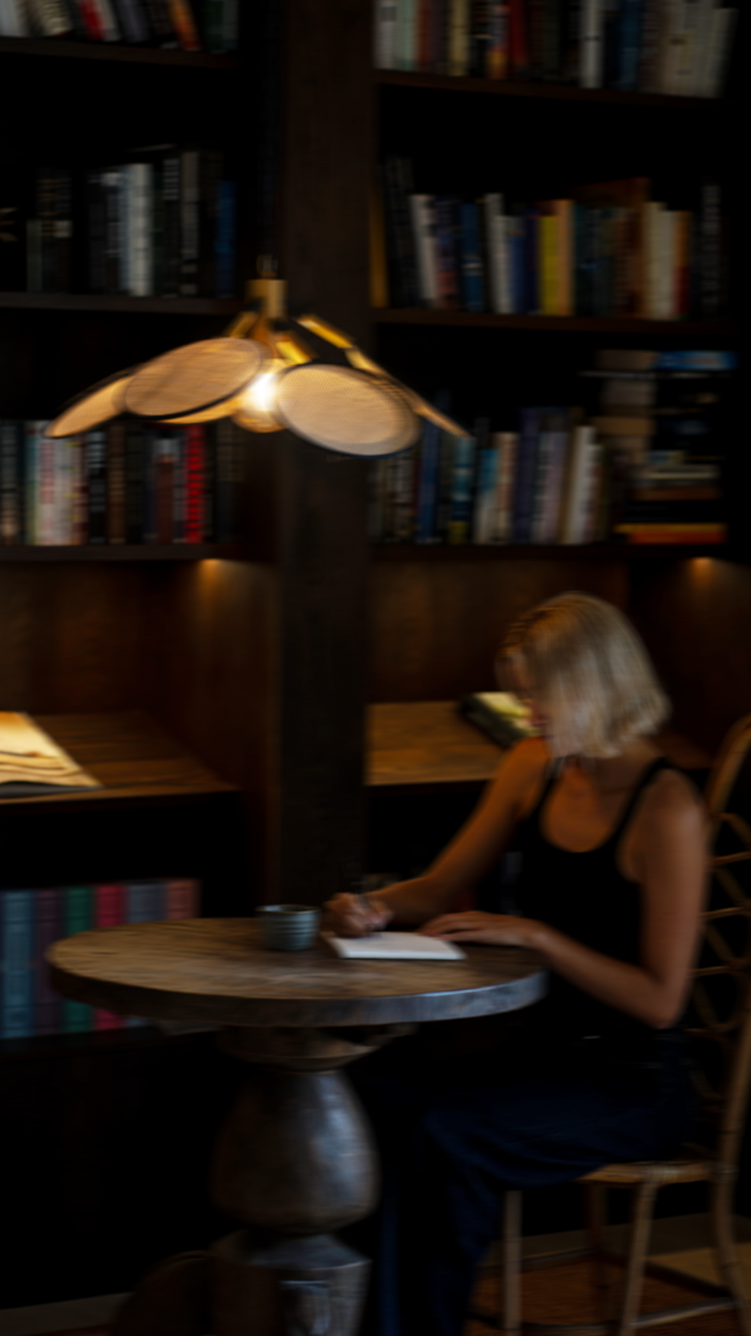 A woman with blonde hair sitting at a round wooden table, writing in a notebook, with bookshelves in the background and a modern hanging lamp above she is in a cozy library or cafe.