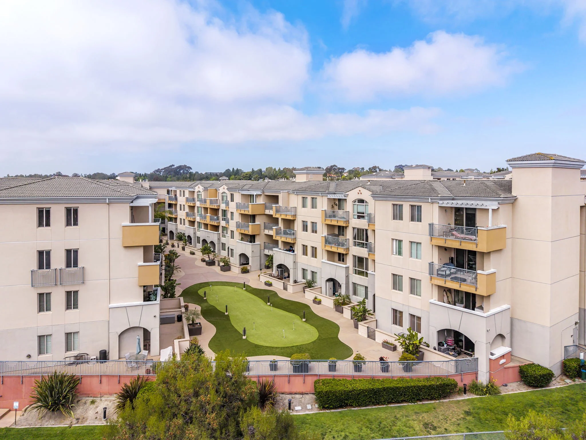 View of a residential apartment complex with multiple beige buildings, balconies, and a landscaped courtyard with green grass, trees, and pathways under a partly cloudy sky.