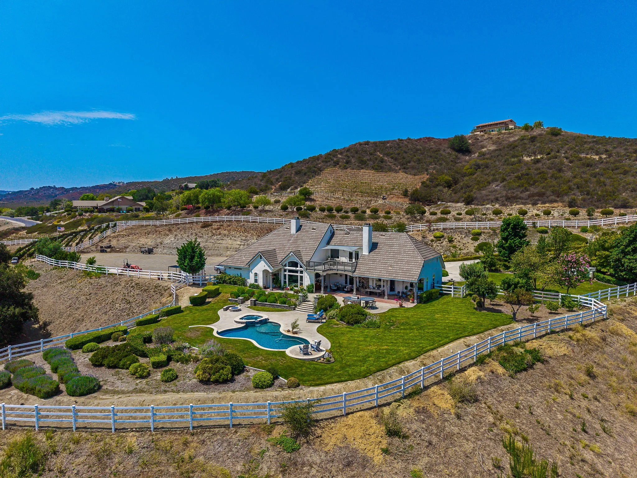 A residential backyard with a swimming pool, landscaped lawn, and a white fence. There are neighboring hills with houses and a clear blue sky.