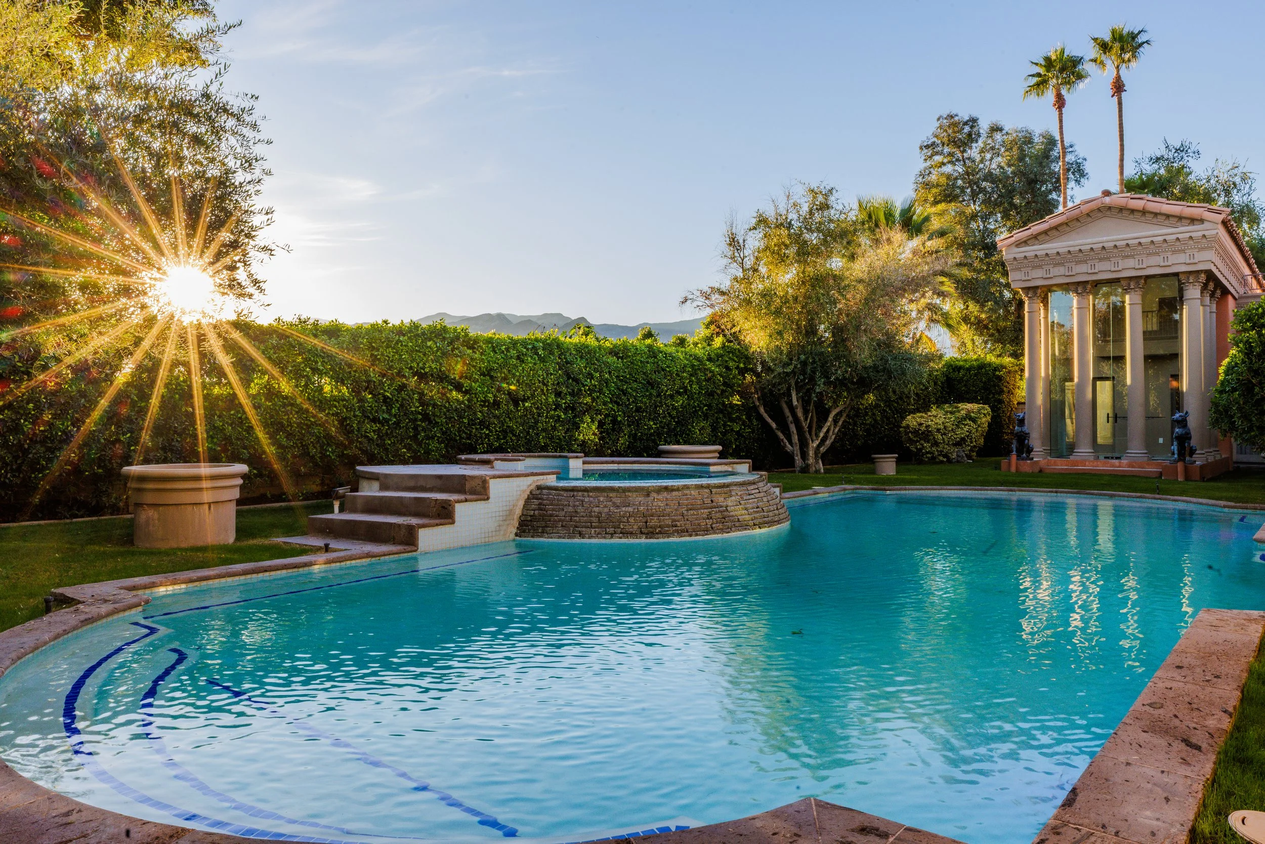 A backyard pool with a curved edge, surrounded by a patio and lush greenery, including trees and bushes, under a clear blue sky with the sun shining brightly.