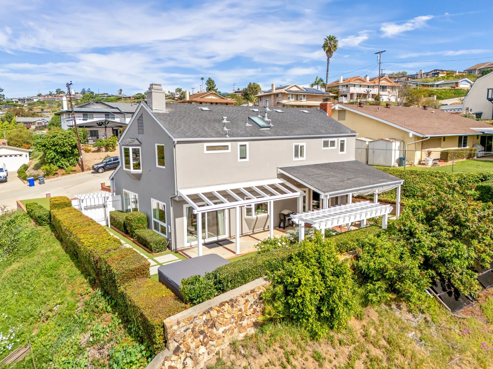 A backyard with a patio and a house with a gray roof, surrounded by lush greenery, with neighboring houses and a hillside in the background under a partly cloudy sky.
