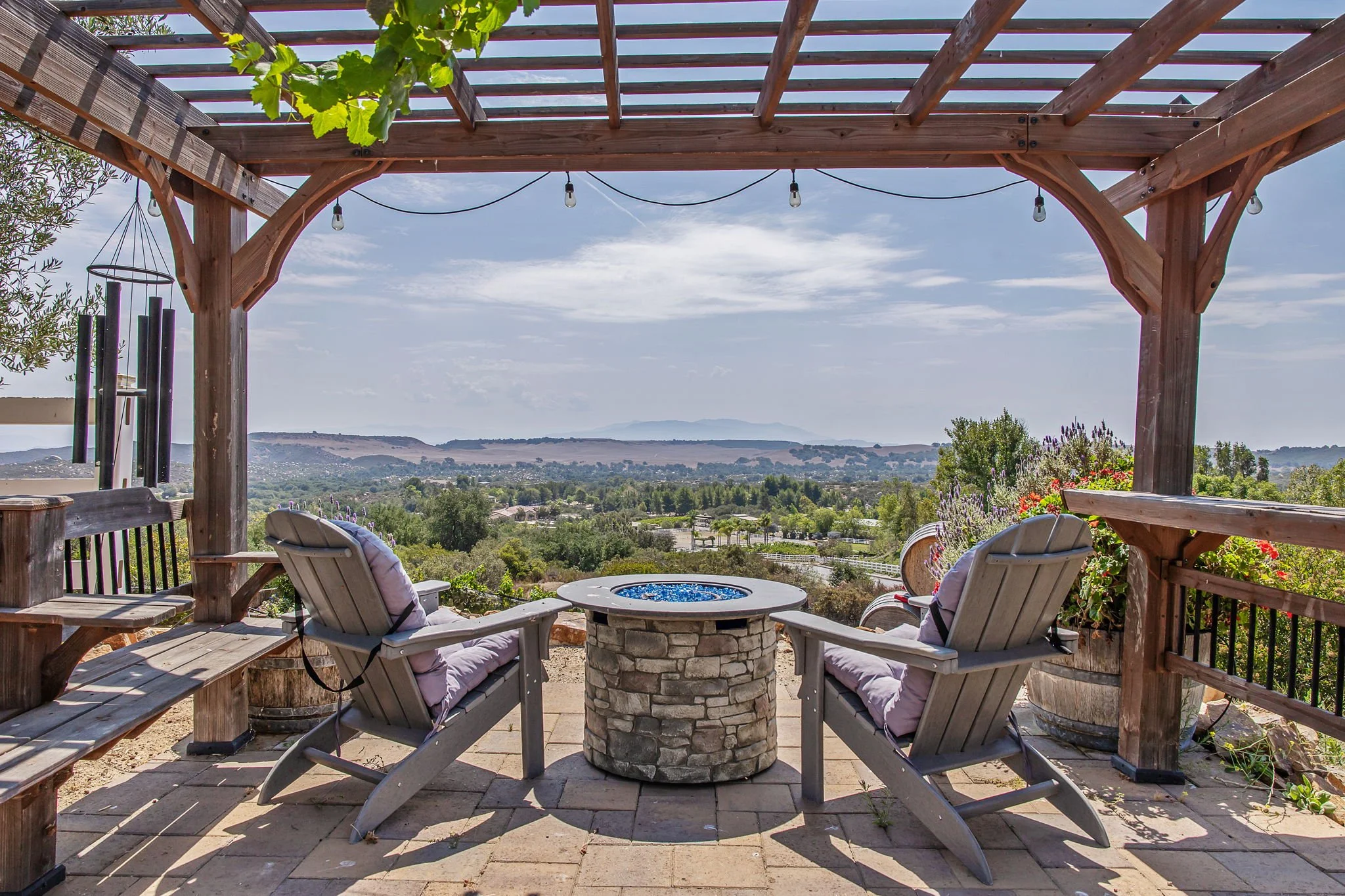 Outdoor seating area with four wooden chairs with cushions, a stone fire pit table with a blue fire feature, under a wooden pergola with string lights, overlooking a scenic landscape with trees and hills in the distance.
