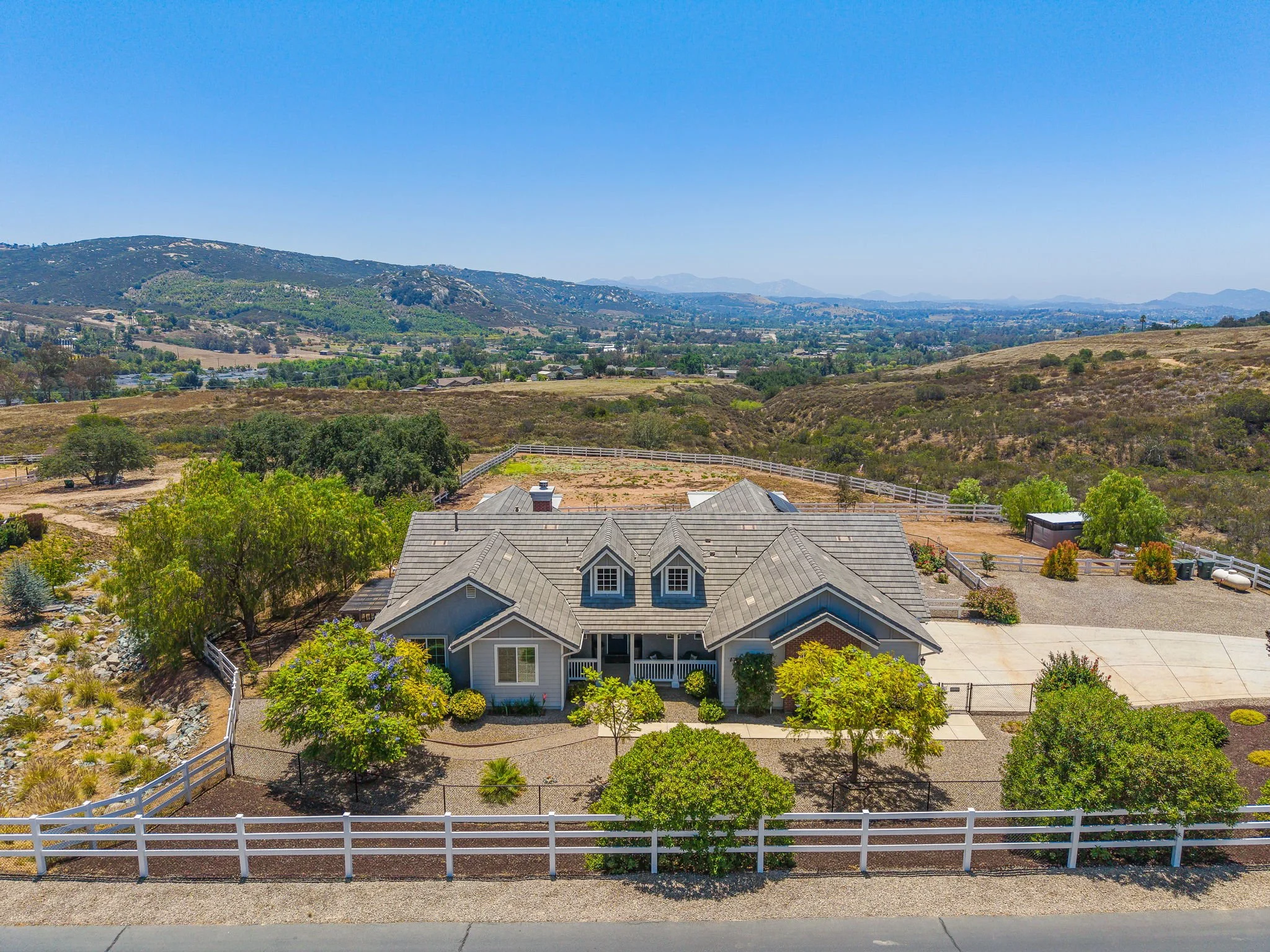 A residential house with gray roof in a green, fenced backyard with trees, bushes, flowers, and a pathway, overlooking rolling hills and distant mountains under a clear blue sky.