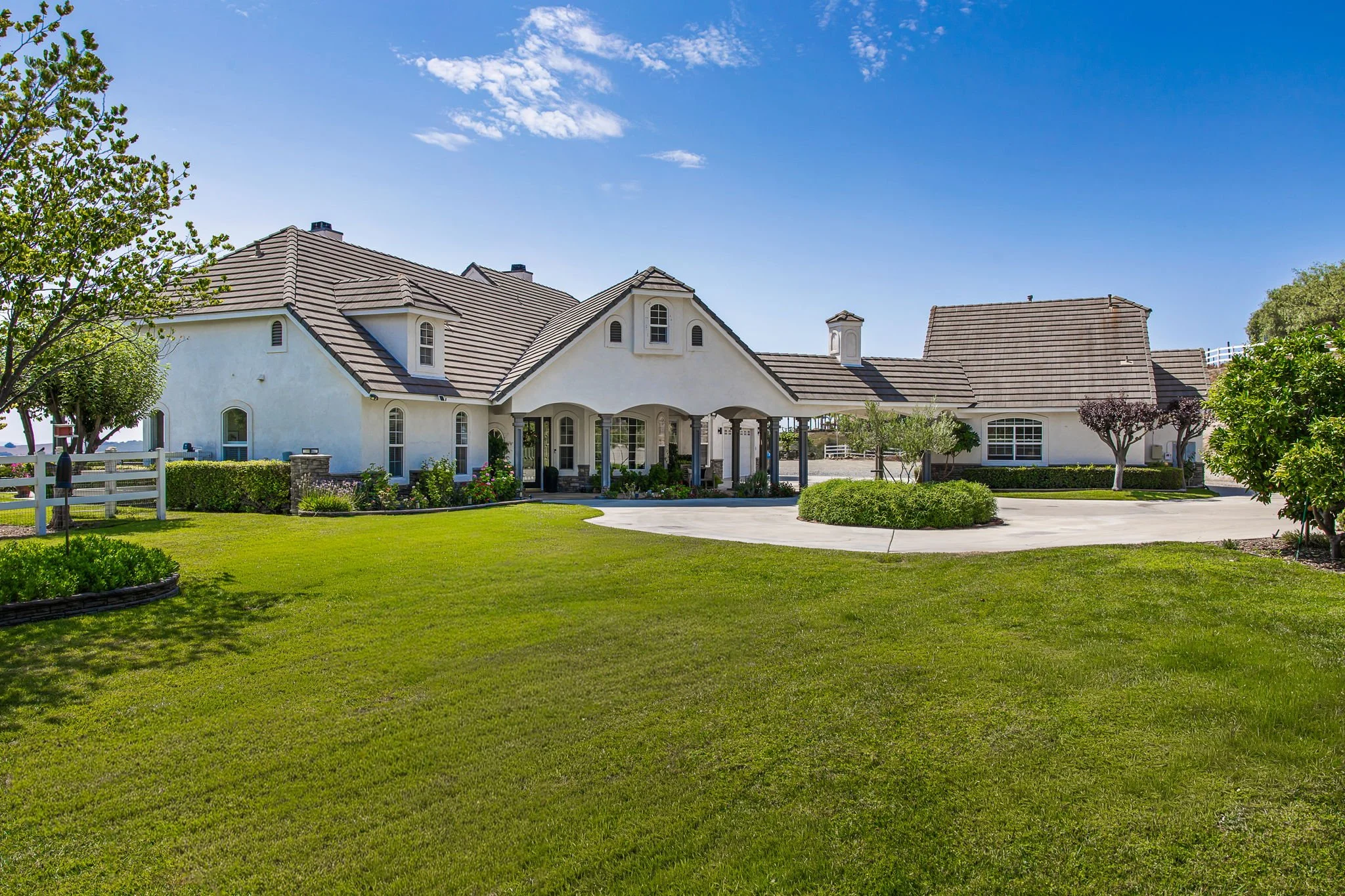 Large white house with a tile roof, front yard with green grass, trees, and bushes, under a bright blue sky with some clouds.