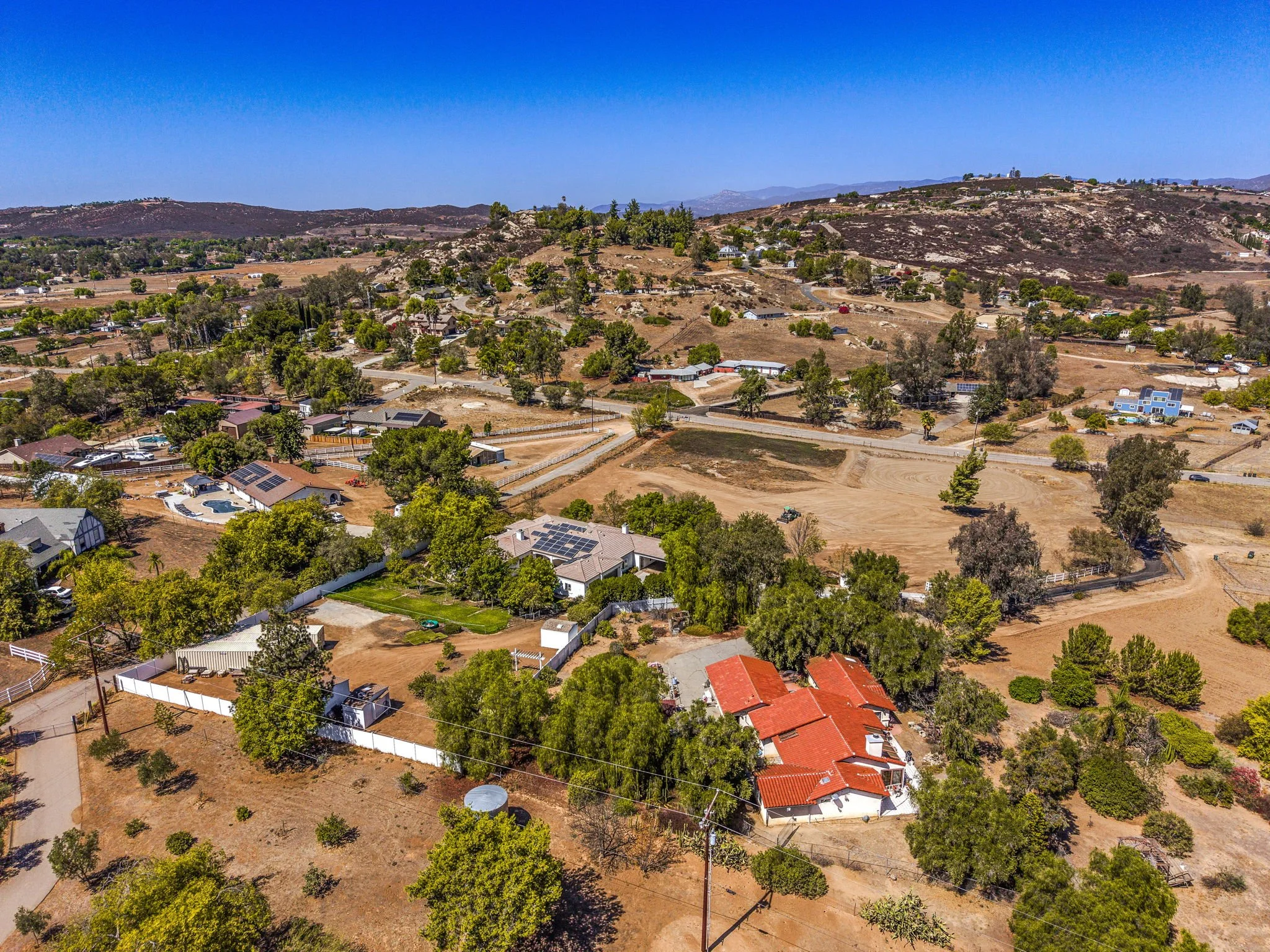 Aerial view of a suburban neighborhood with houses, trees, and dirt roads on a hillside under a clear blue sky.