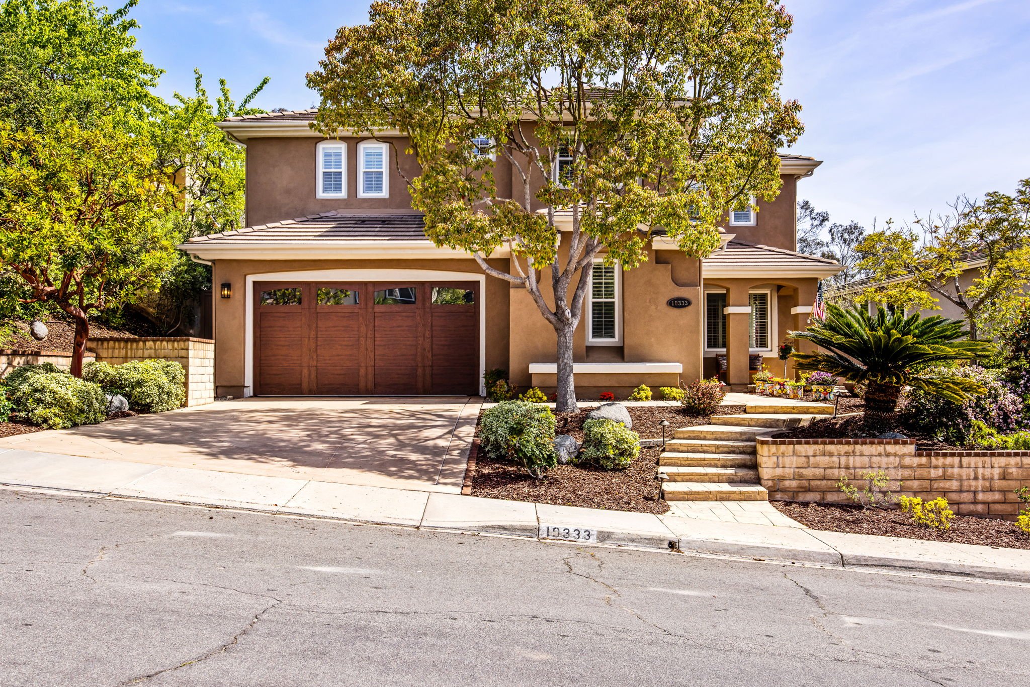 A two-story beige house with a brown garage door, surrounded by green landscaping and trees, with a sidewalk and curb marked 19333 in front.